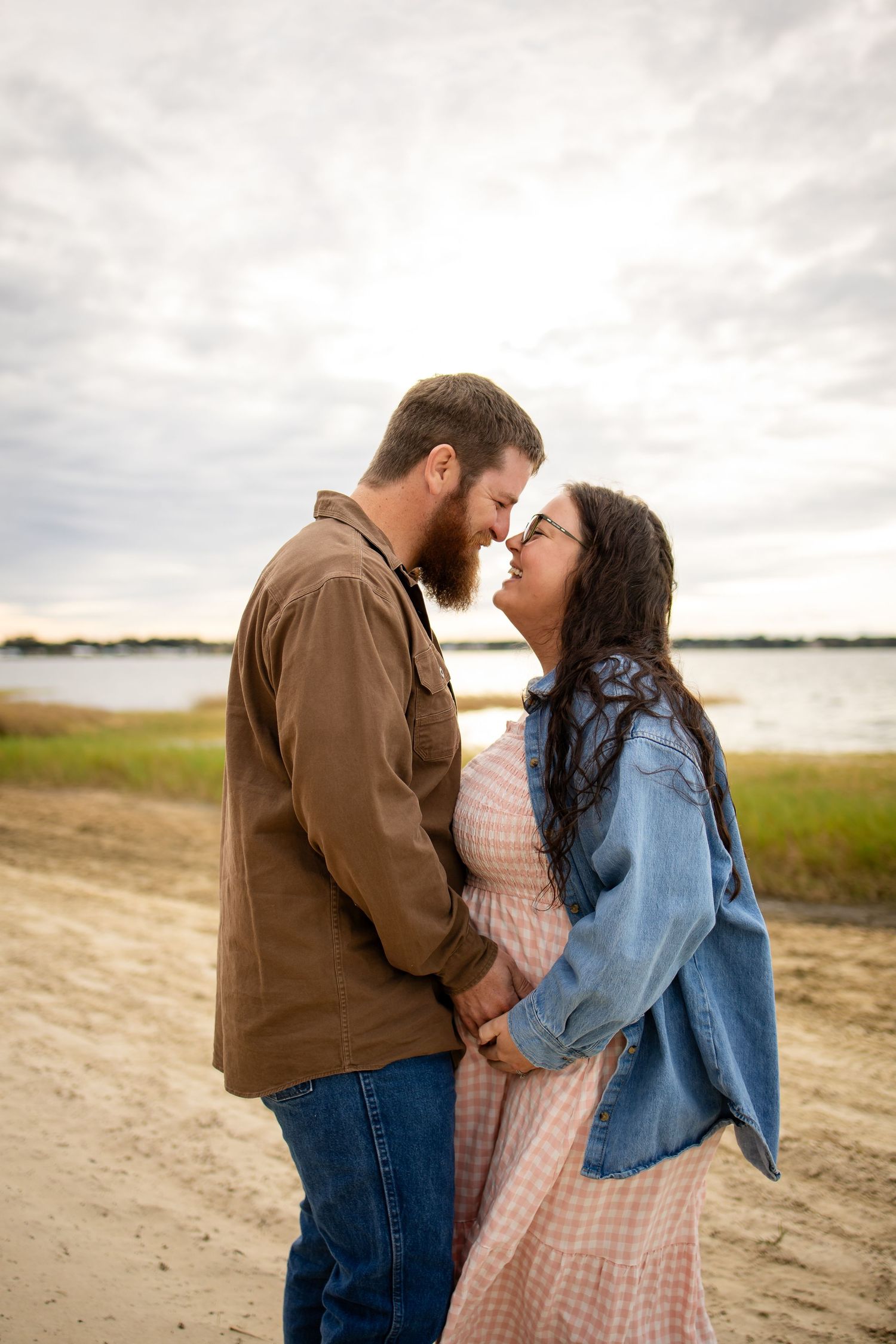 A couple embraces tenderly against an overcast sky with marshland and water visible in the distance.