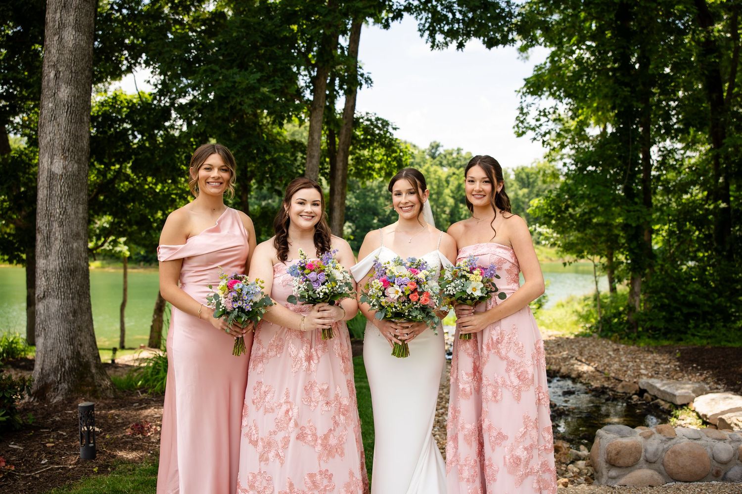 Bride and three bridesmaids in pink floral dresses holding colorful bouquets outdoors in a lush green wooded setting.