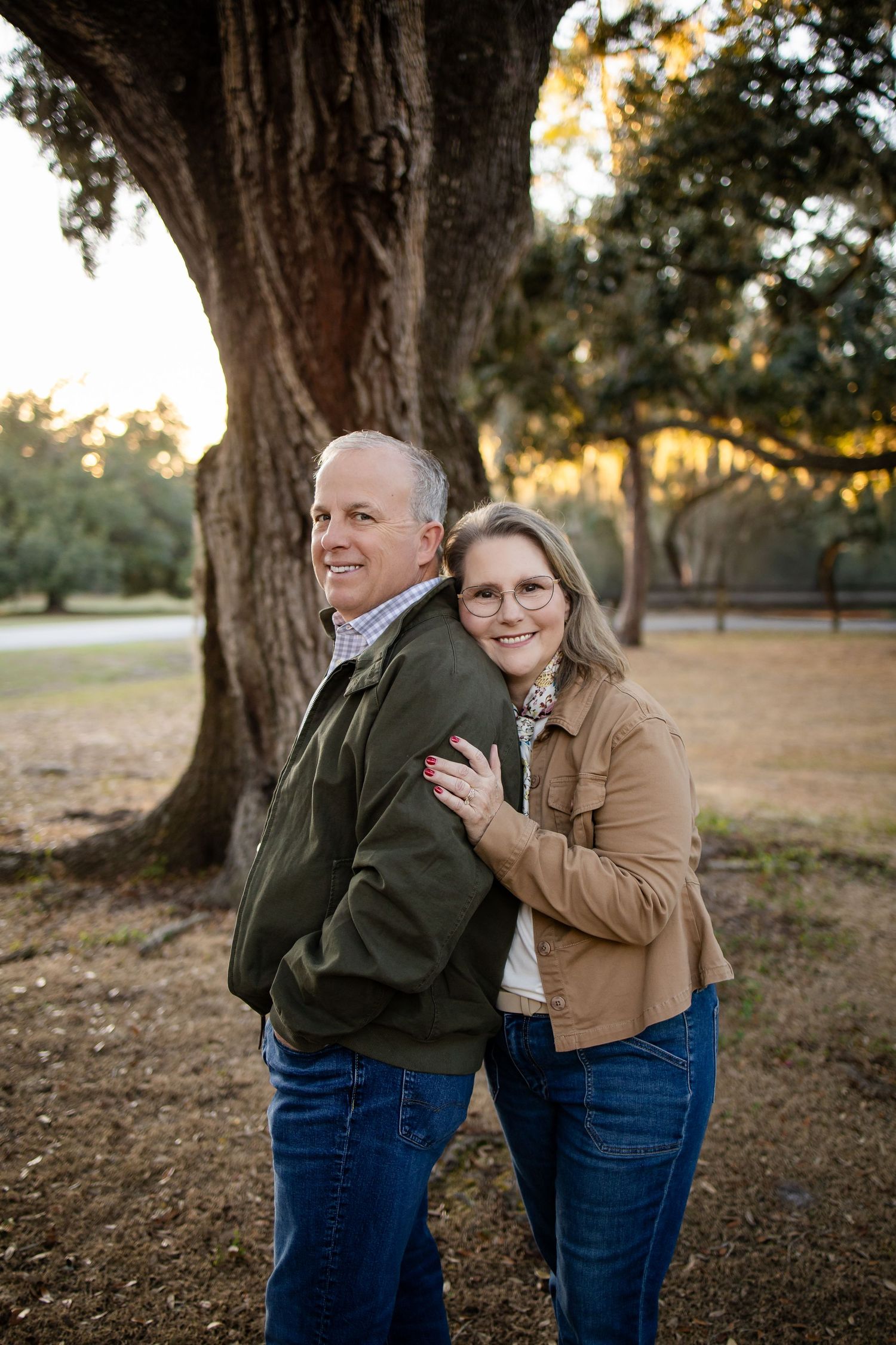 A couple shares candid moments while posing for photos under large oak trees at dusk.