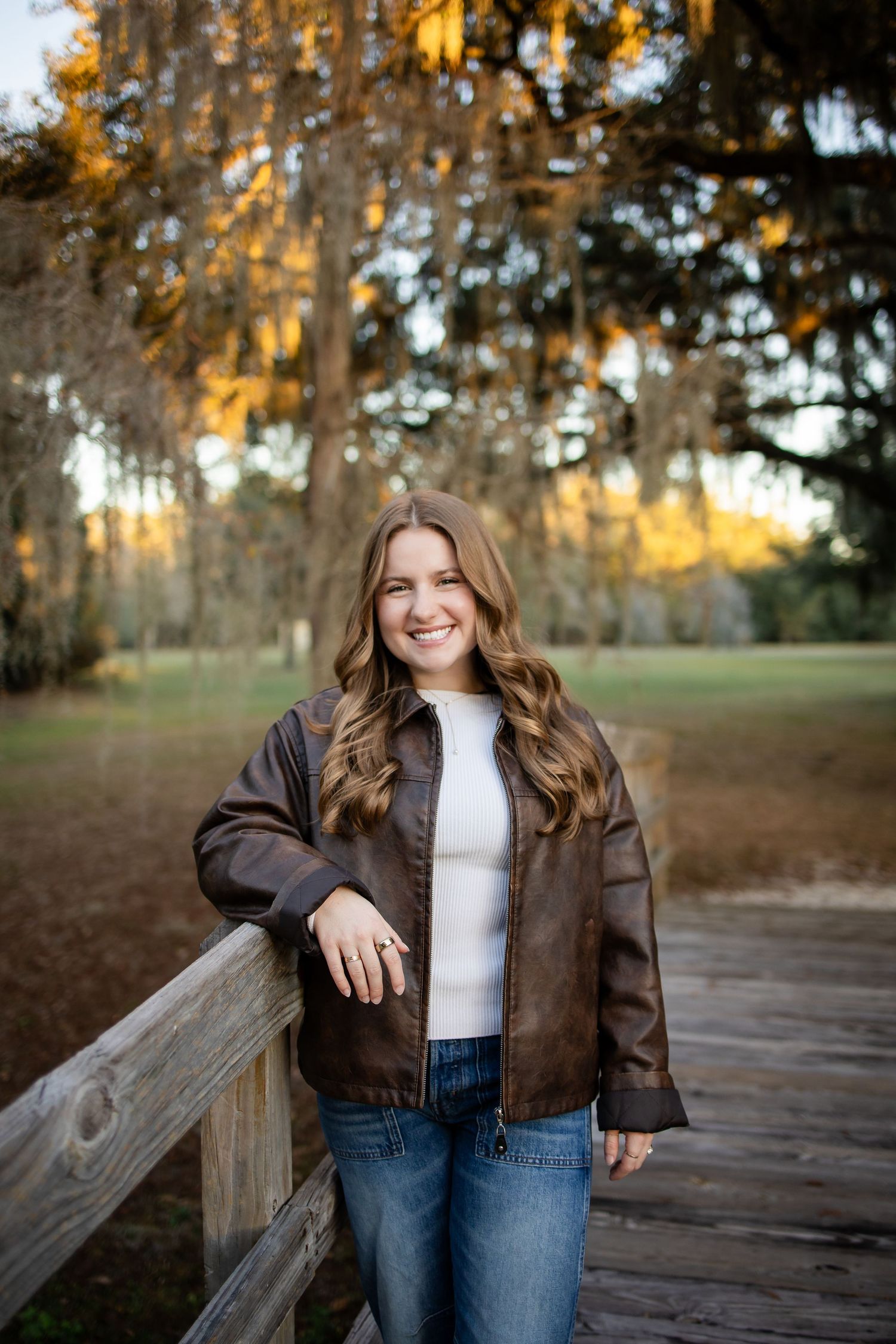 Person in brown leather jacket and jeans poses casually on wooden bridge during sunset.