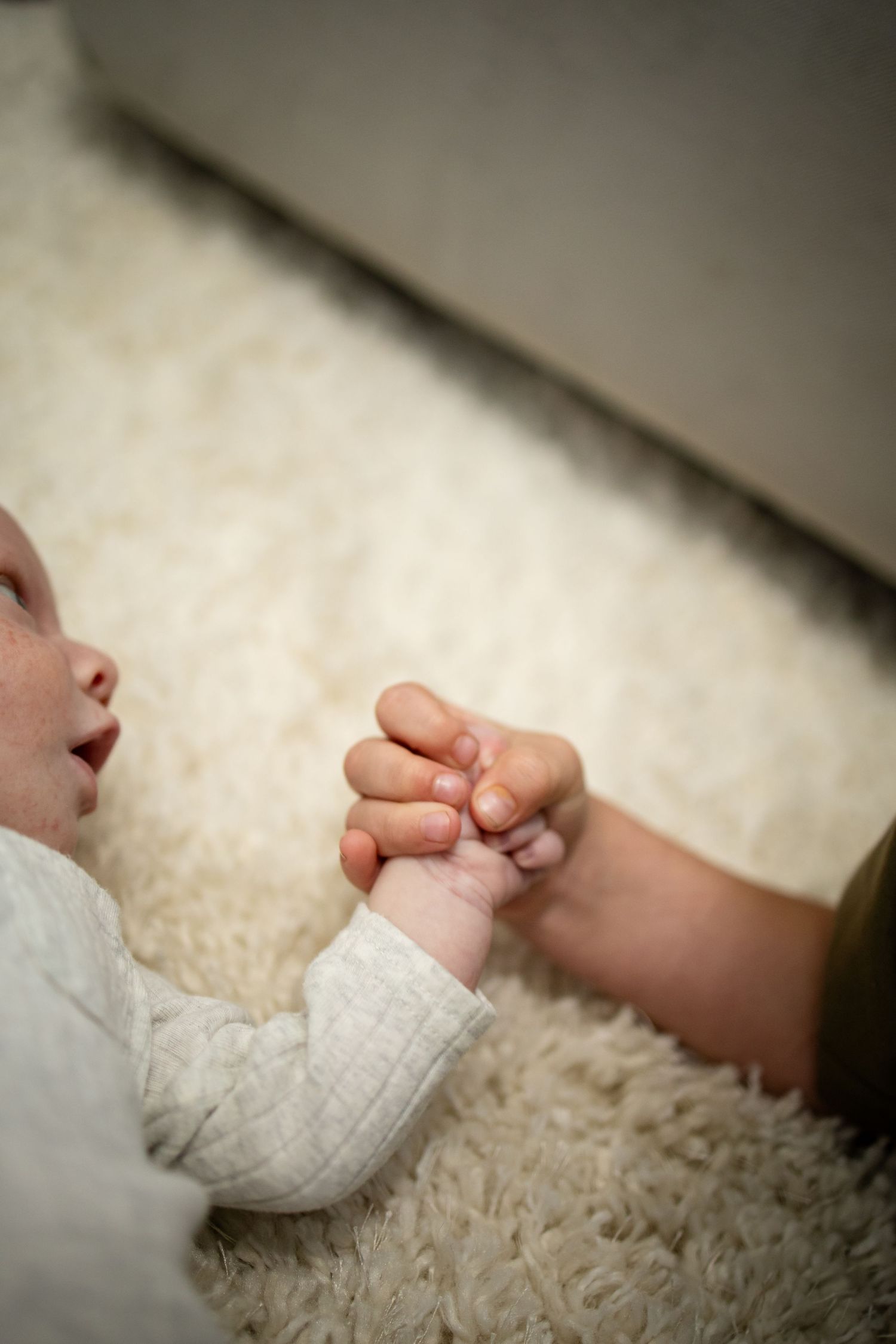 A tender moment as a baby's small hand grasps an adult's fingers against a soft white carpet background.
