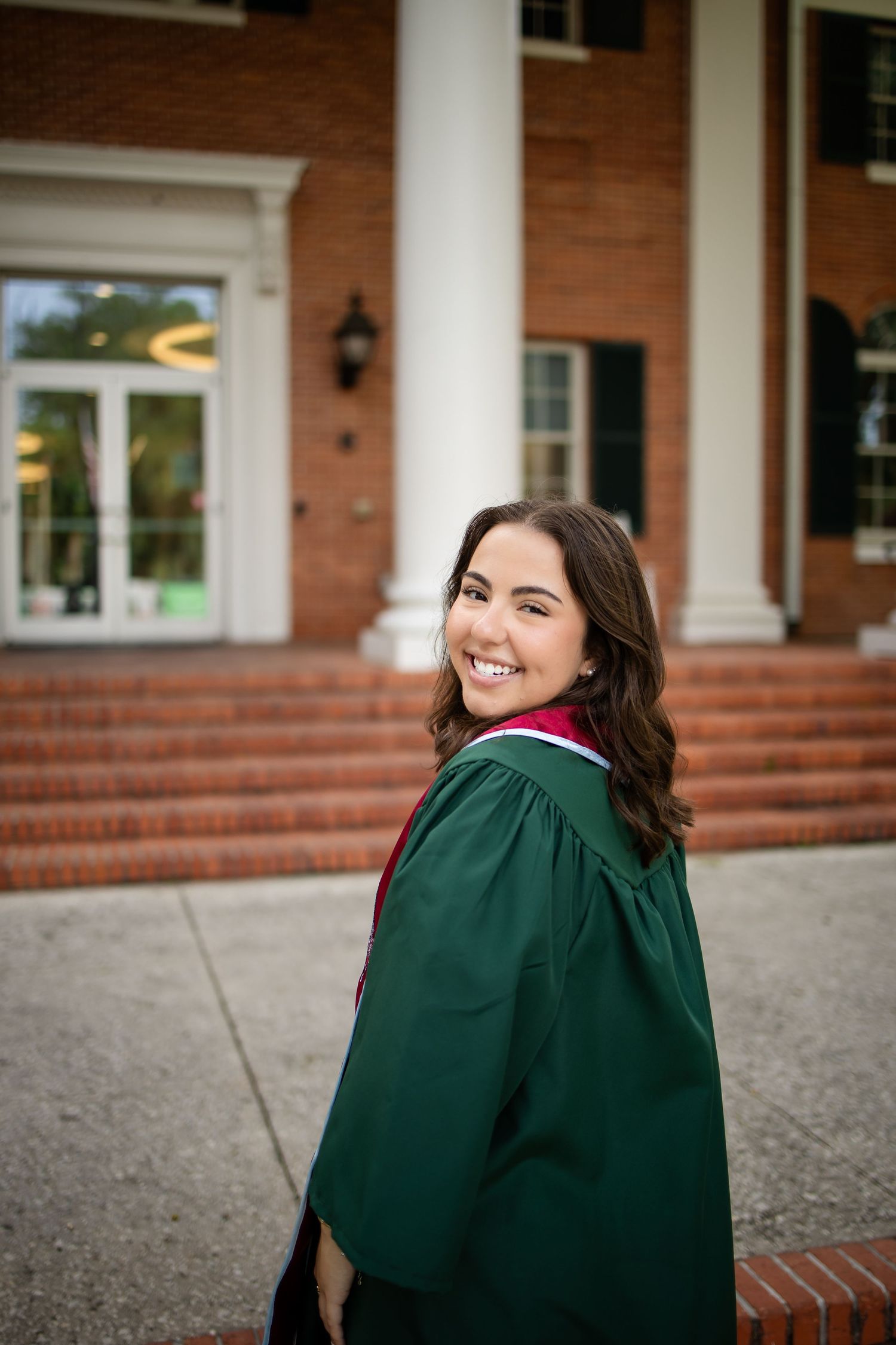 Graduate in a green academic gown poses with a bright smile in front of a brick building with white columns.