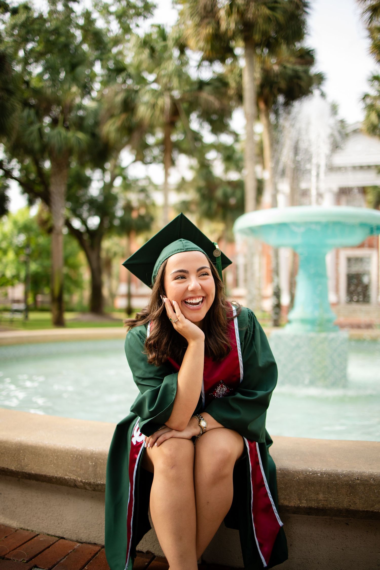 Graduate in green cap and gown sitting by ornate fountain on sunny campus day.