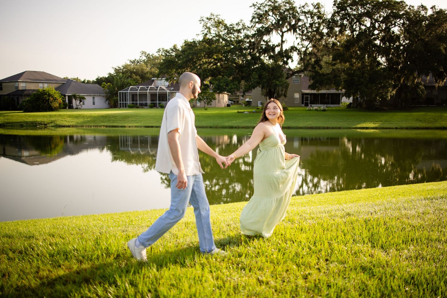 Two figures walk together and pose playfully at sunset by a serene pond surrounded by green grass and trees.