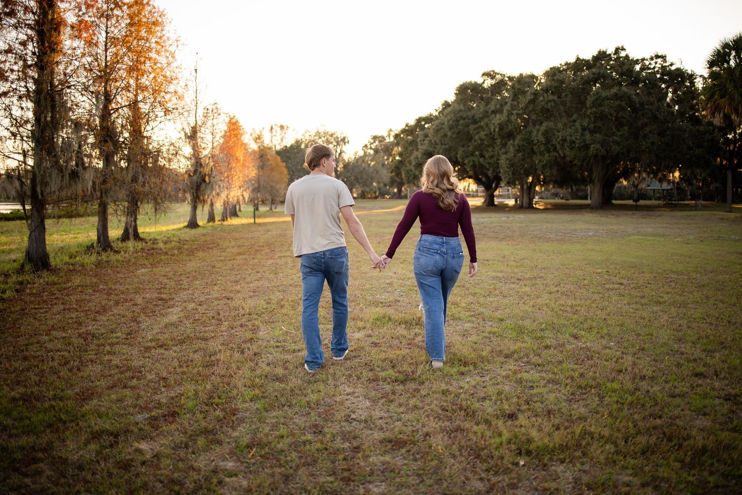 A couple walks hand in hand through a sunlit autumn field at golden hour.