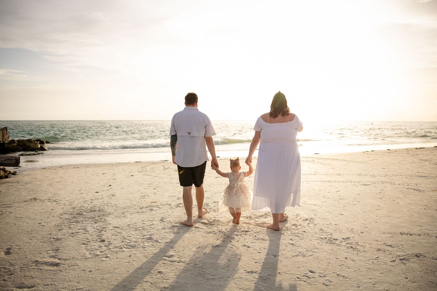 Family of three walking hand-in-hand on a sandy beach at sunset, parents guiding toddler toward ocean waves.