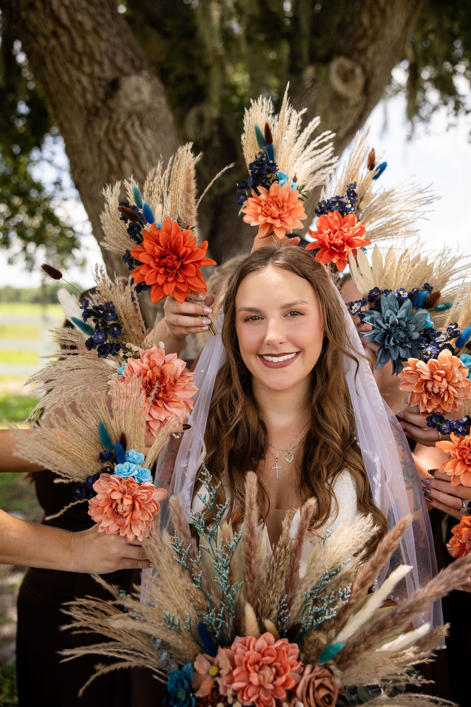Smiling bride surrounded by colorful boho bouquets with orange, teal, and dried pampas grass flowers outdoors.