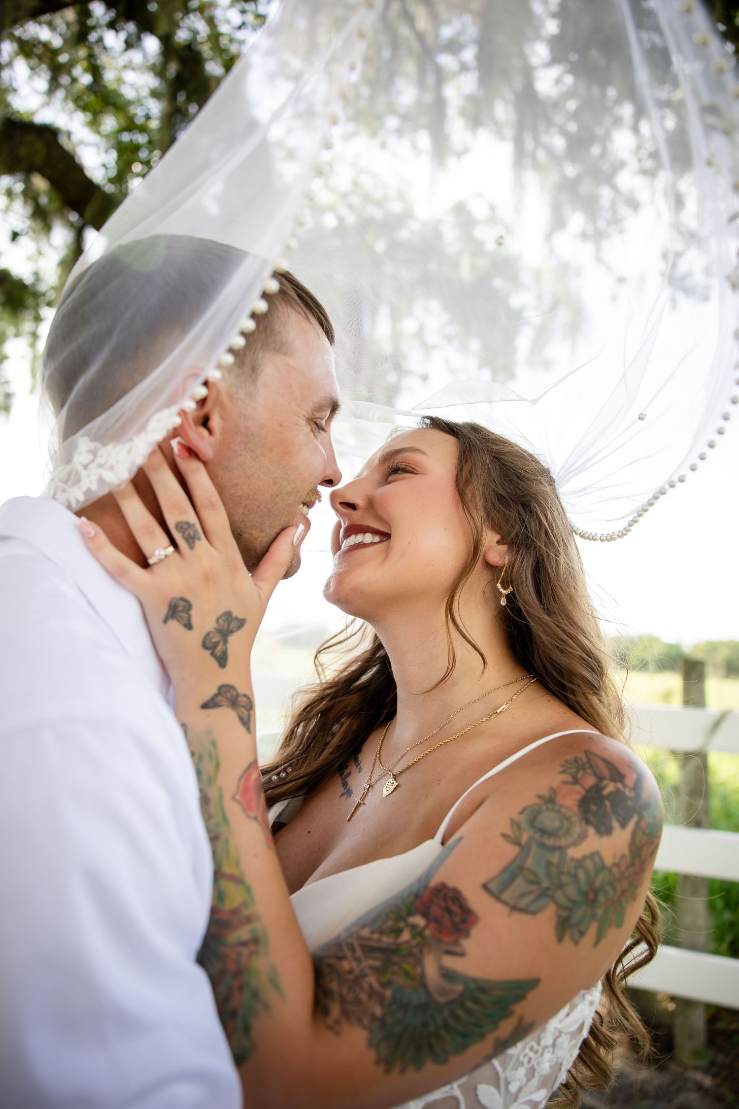 Bride and groom share a romantic moment under a wedding veil, both covered in tattoos, smiling at each other outdoors.