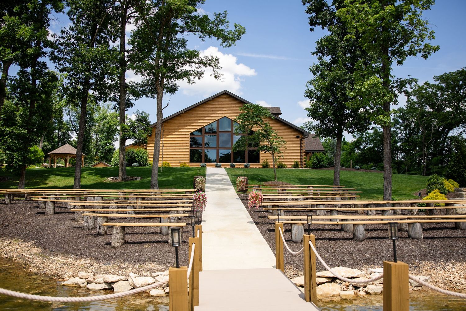 Outdoor wedding venue with wooden chapel, pew seating, and walkway surrounded by lush trees and manicured grounds.
