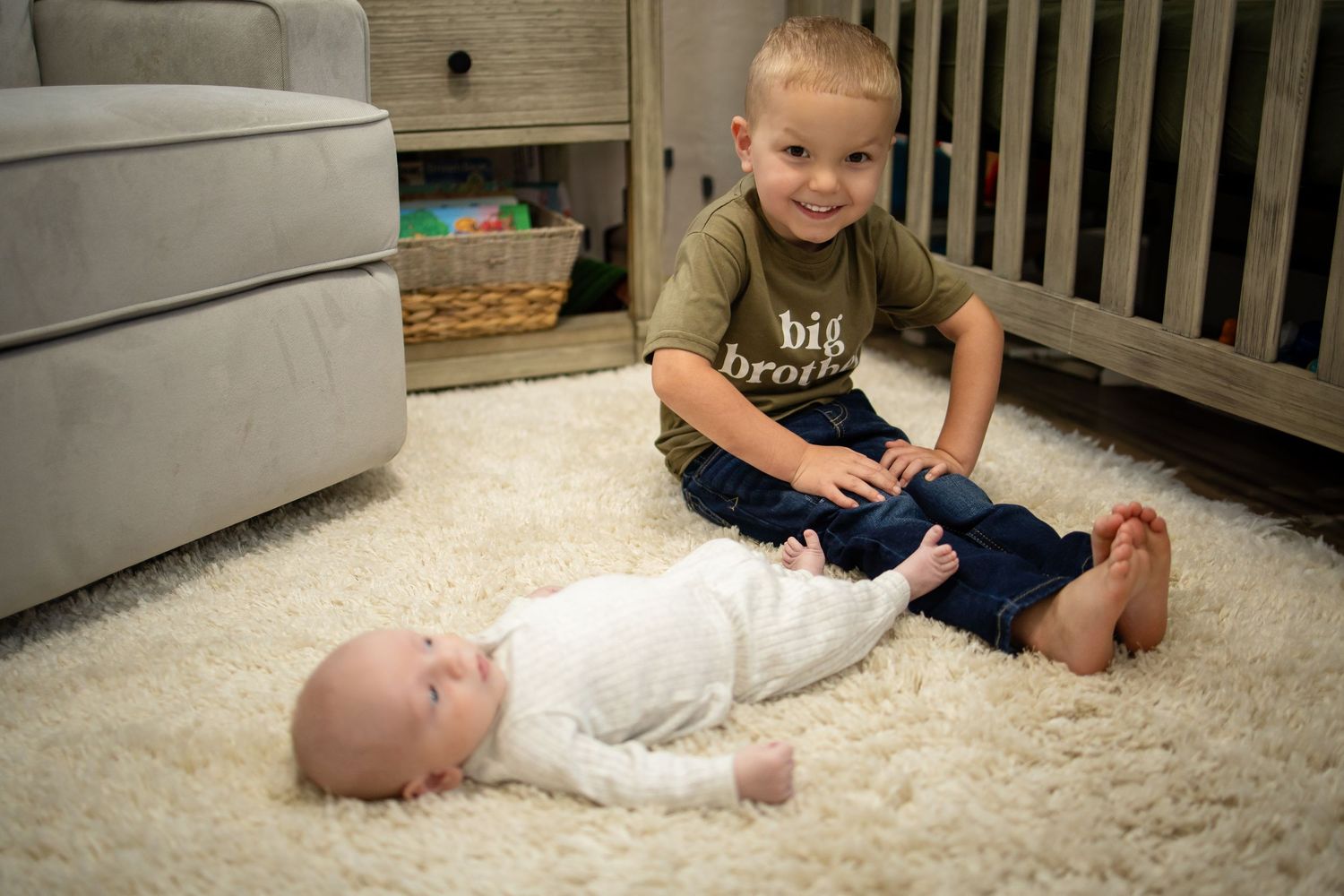Two children sitting together on a white carpet in a living room setting with furniture visible in the background.