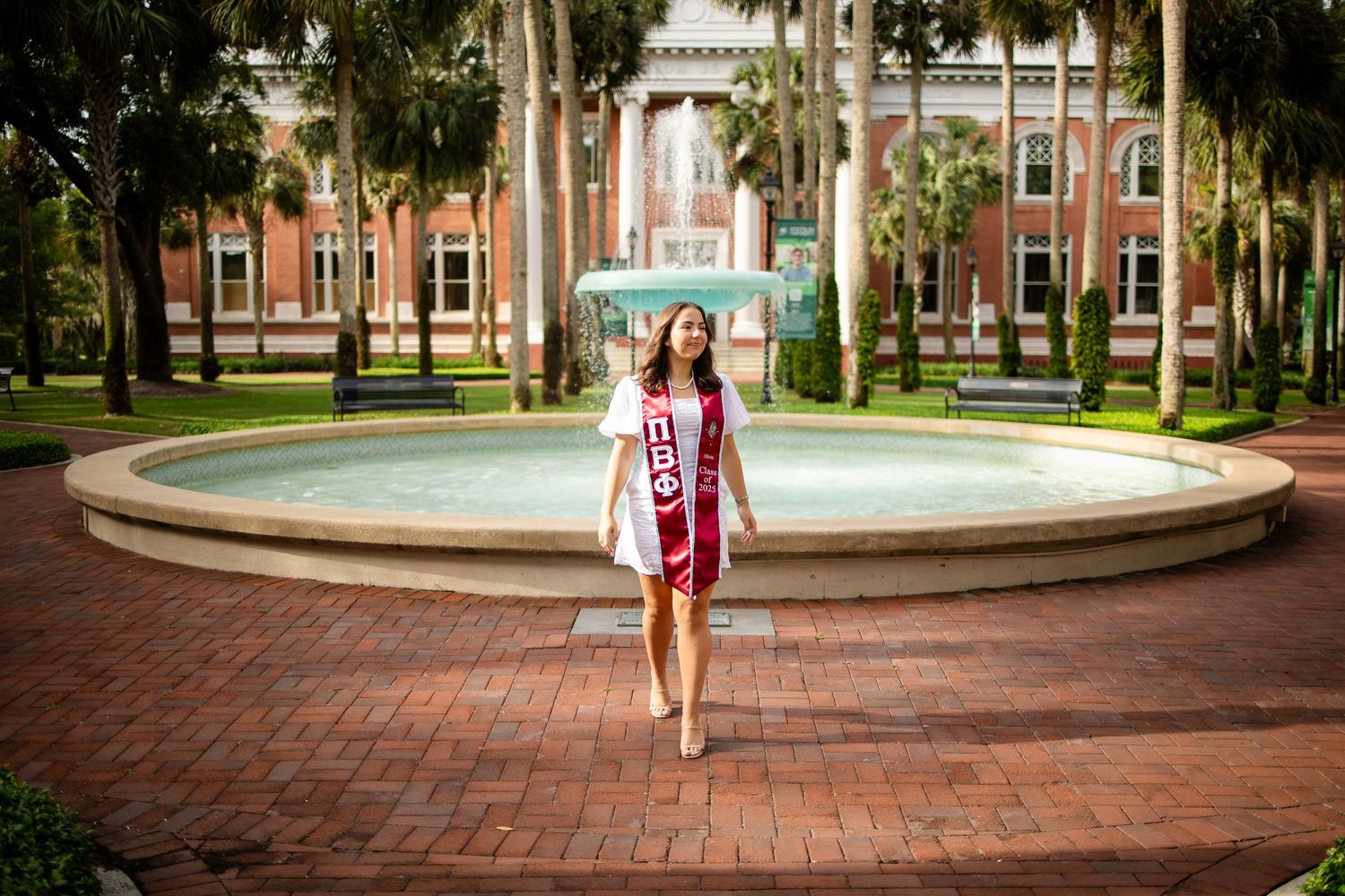 Graduate stands in front of brick fountain at university campus surrounded by palm trees and historic buildings.
