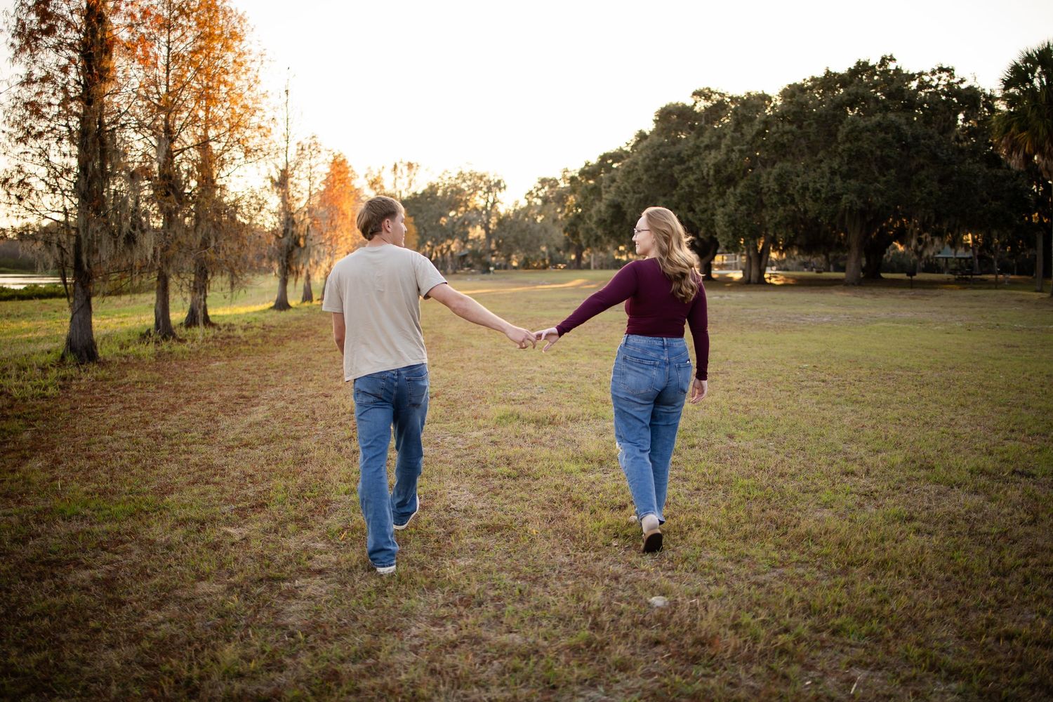 A couple walks hand in hand through a grassy field at sunset with autumn trees in the background.