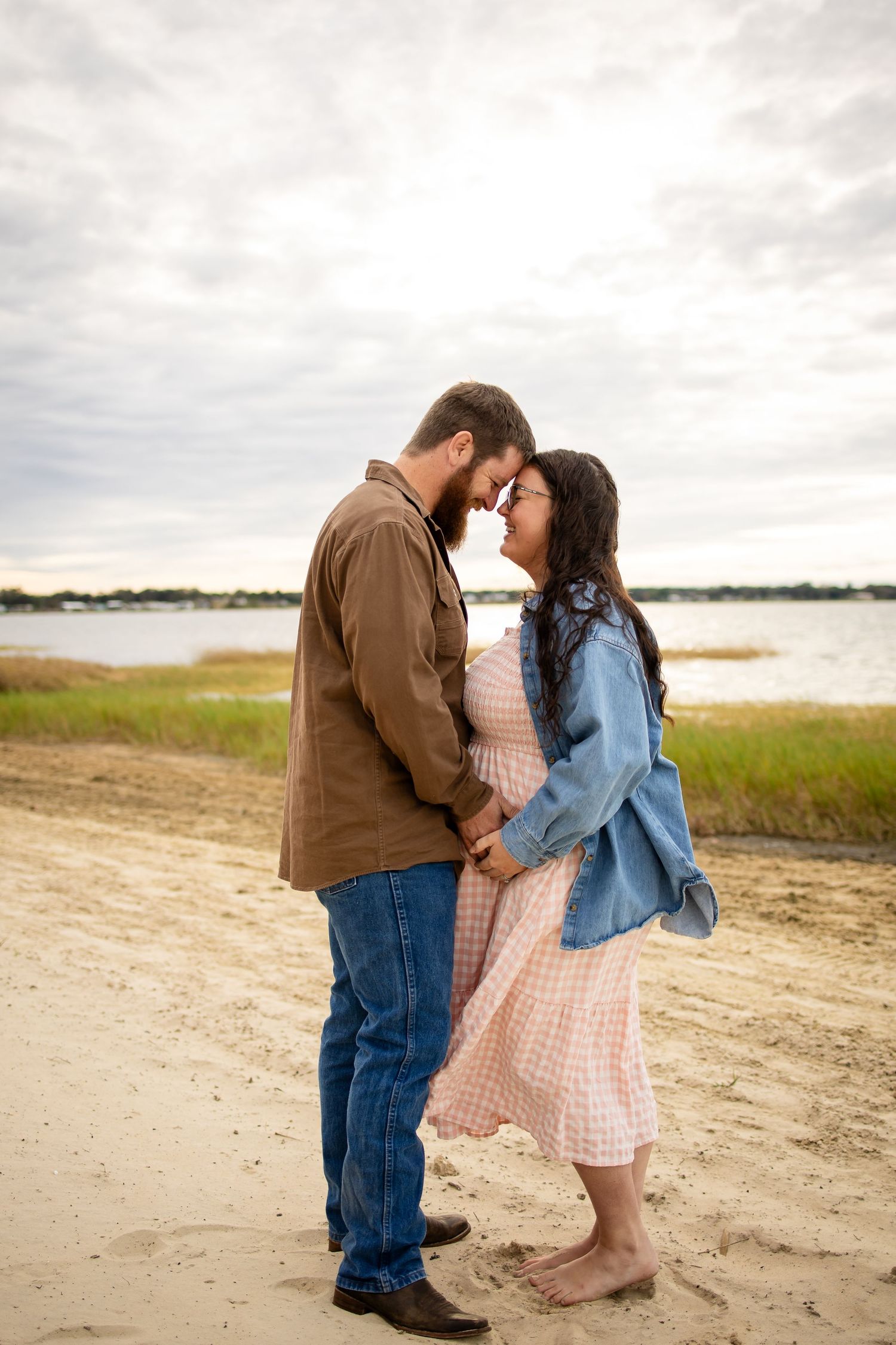 Romantic couple embraces on sandy beach with water and cloudy sky in background.