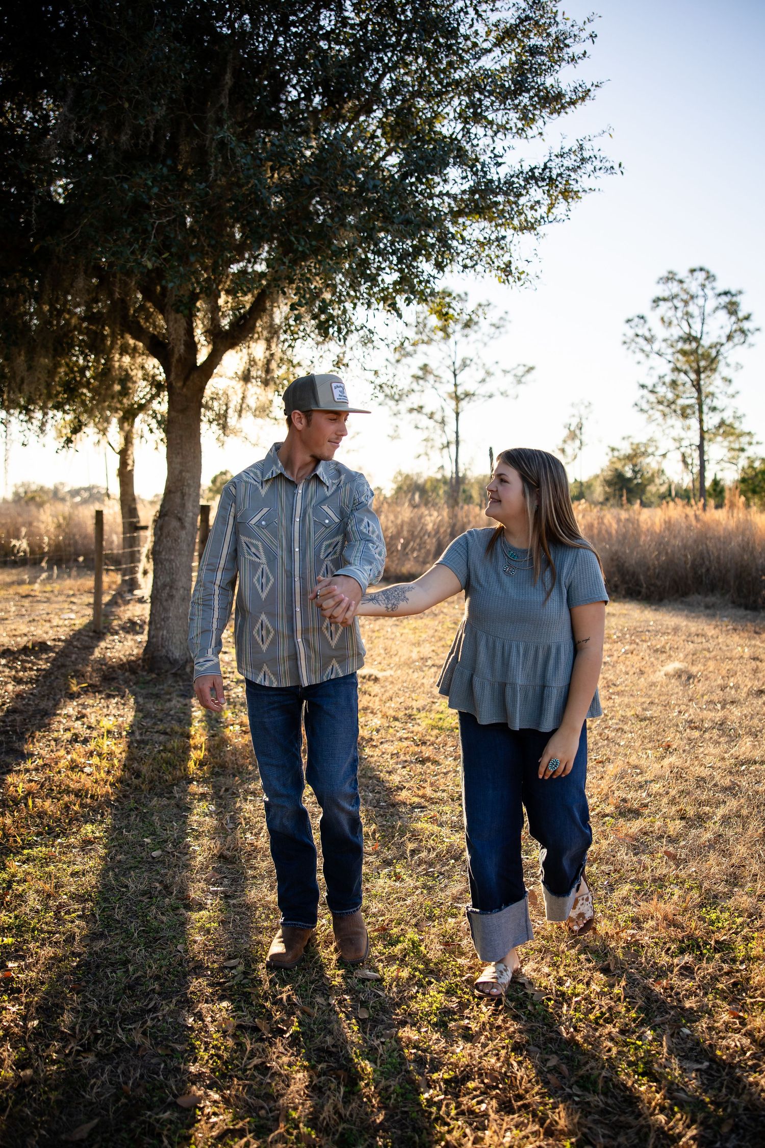 A couple shares a tender moment in a sunlit field with tall pine trees casting long shadows at golden hour.