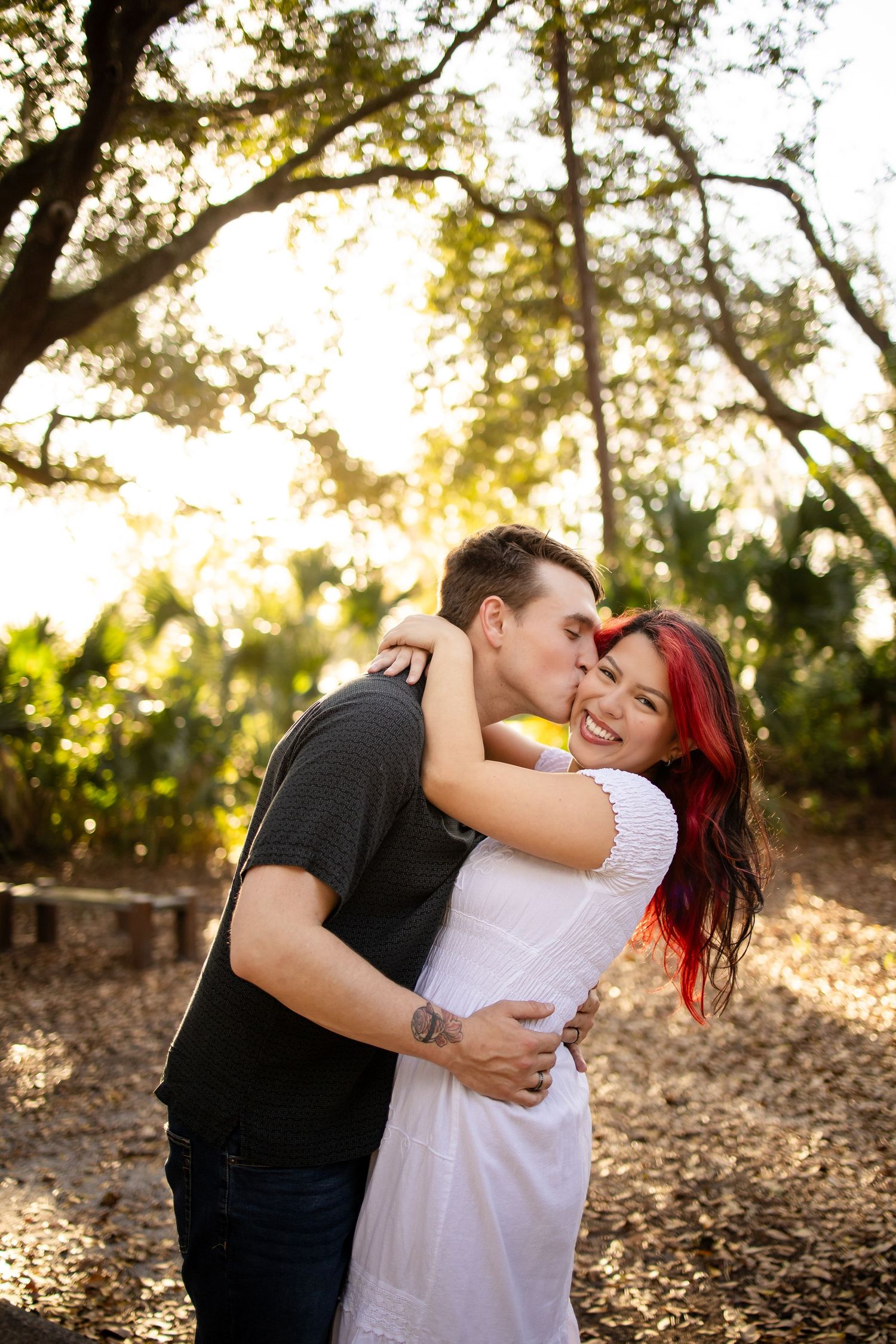 A couple embraces outdoors in a wooded area during golden hour, surrounded by fallen leaves and dappled sunlight.