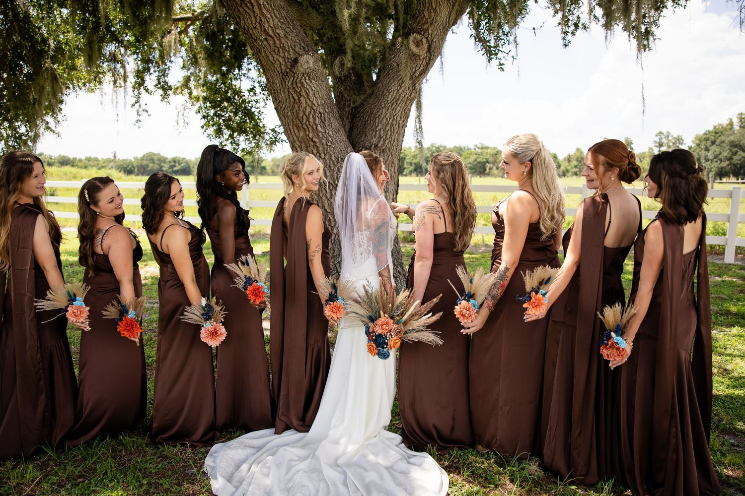 Bride in white gown and veil stands with bridesmaids in brown dresses holding colorful bouquets under a large oak tree.