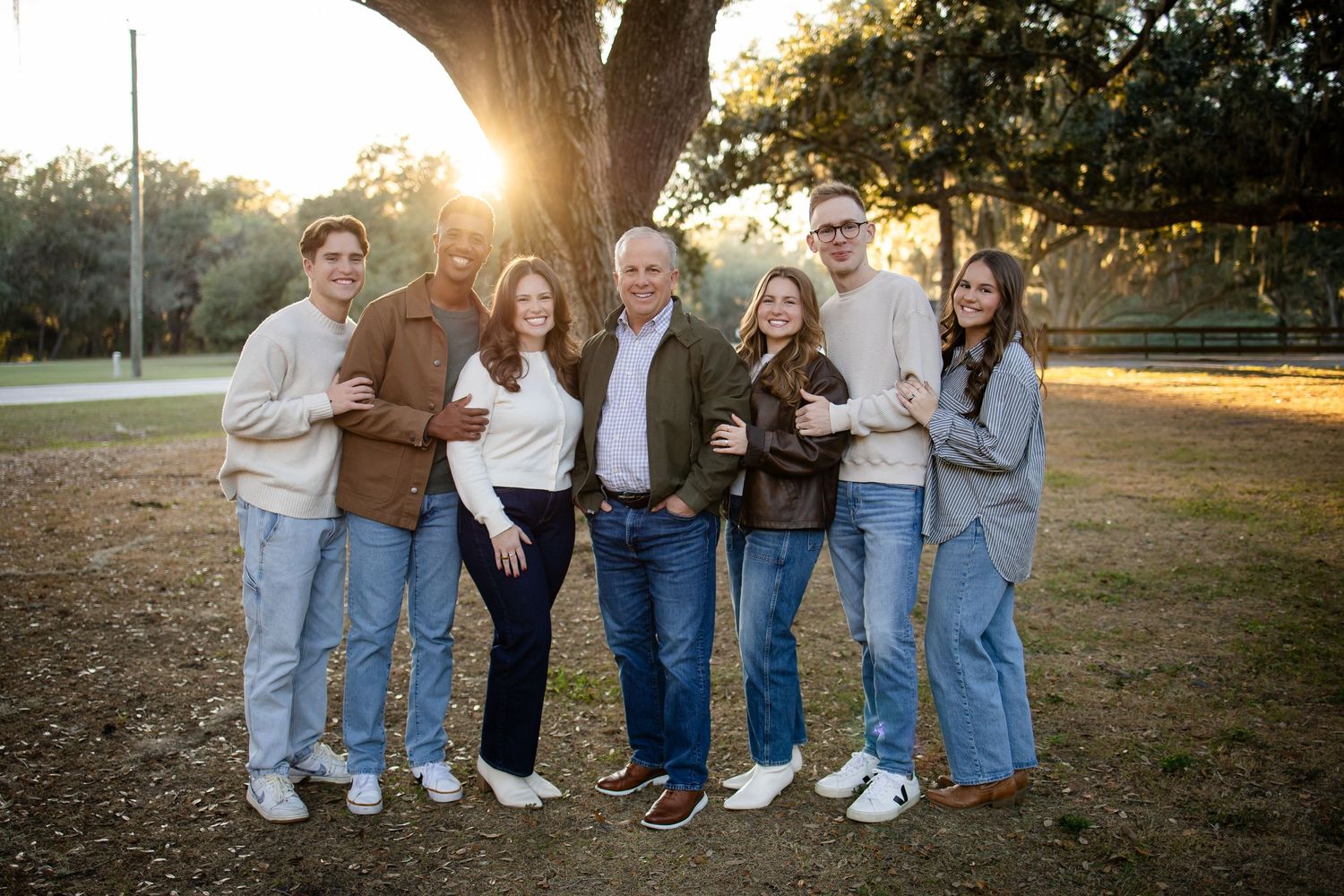 Family group wearing matching casual outfits stands together under sprawling oak tree.