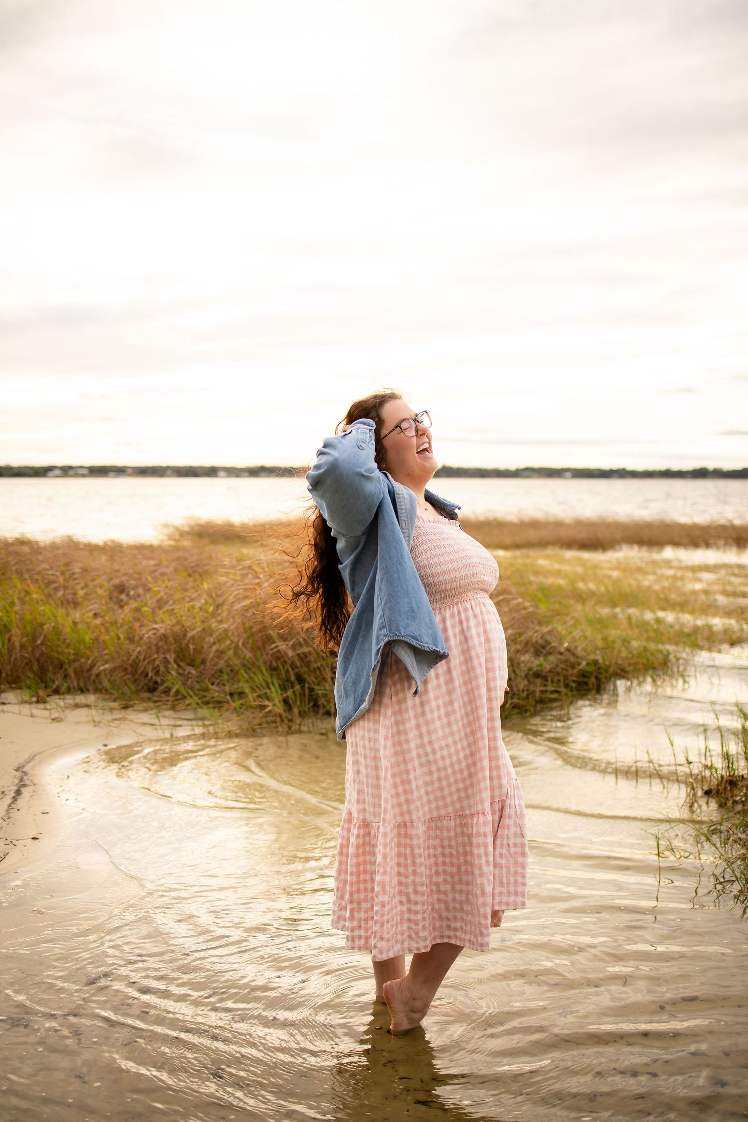 A pregnant woman in a pink gingham dress stands in shallow water at the beach with windswept hair.