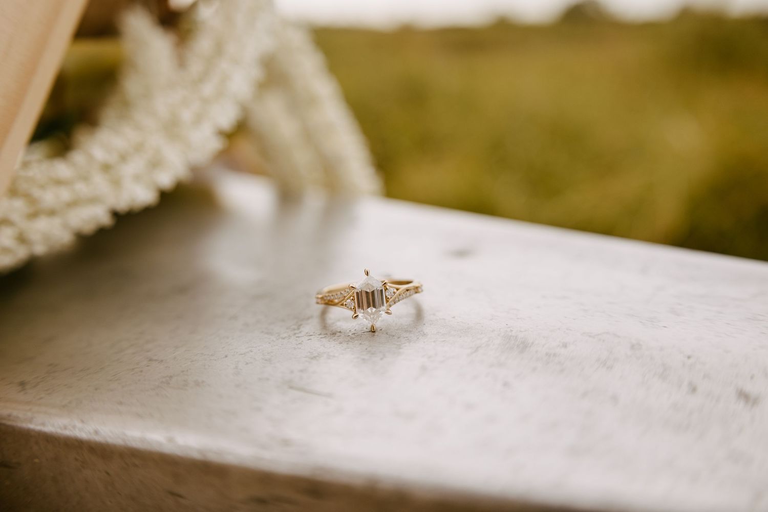 A dainty diamond engagement ring sits on a white surface with a blurred warm background.