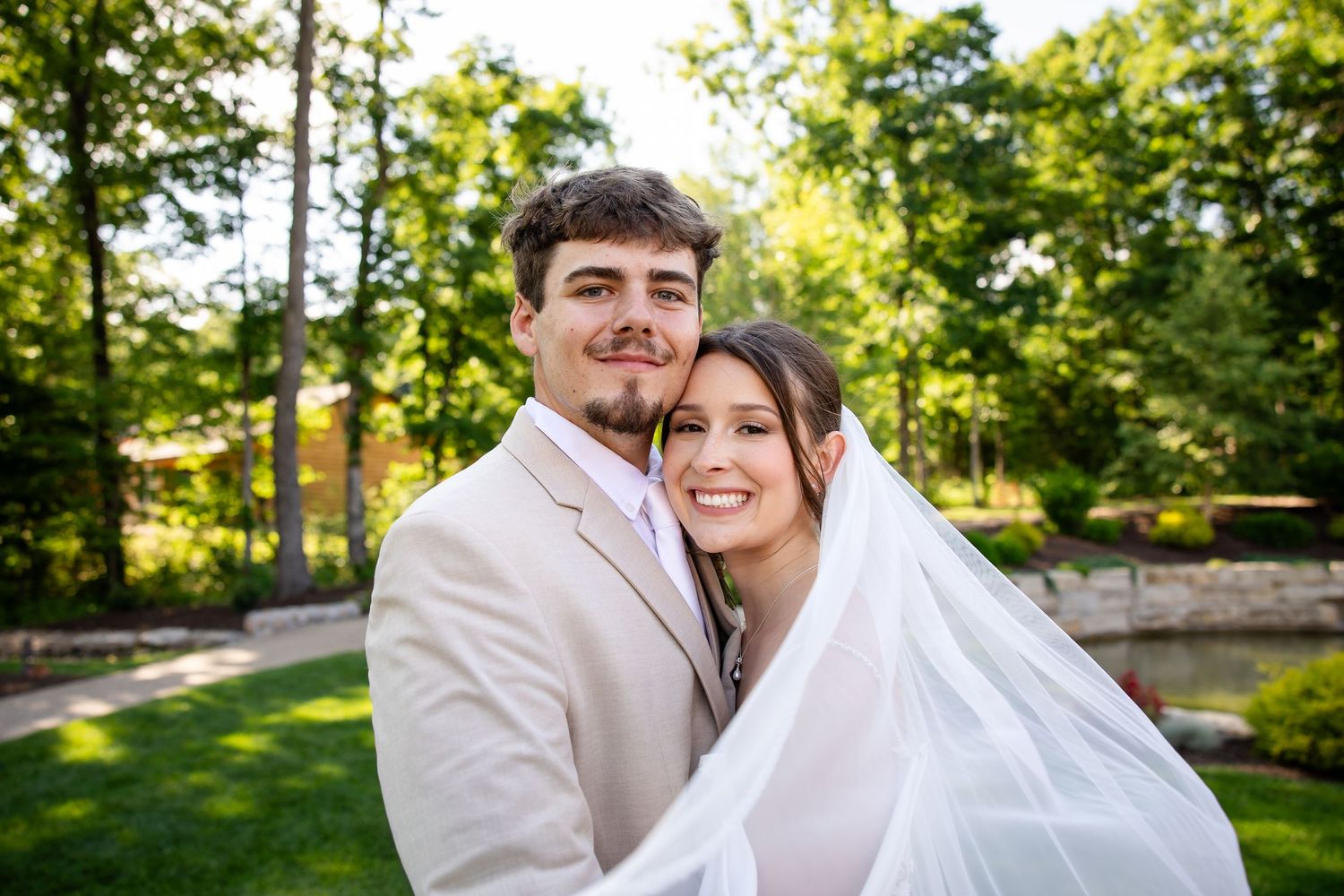 Bride and groom smiling together outdoors in lush green garden setting, groom in tan suit, bride in white veil.