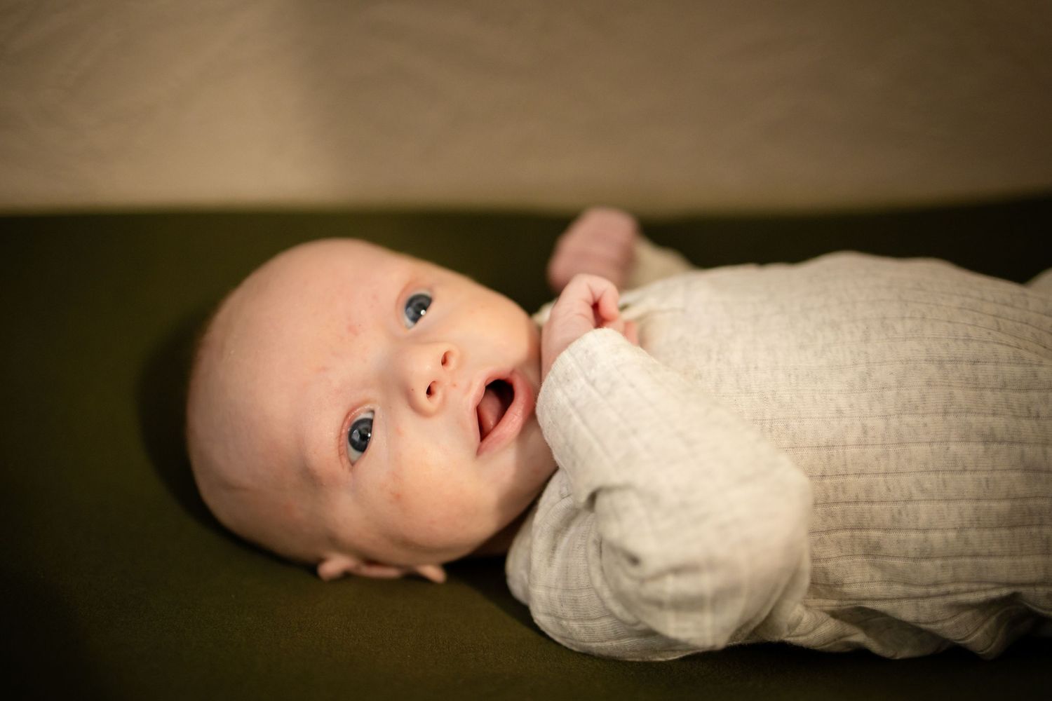 A baby wearing a cream-colored sweater lies on a dark couch while looking up with bright eyes under soft lighting.