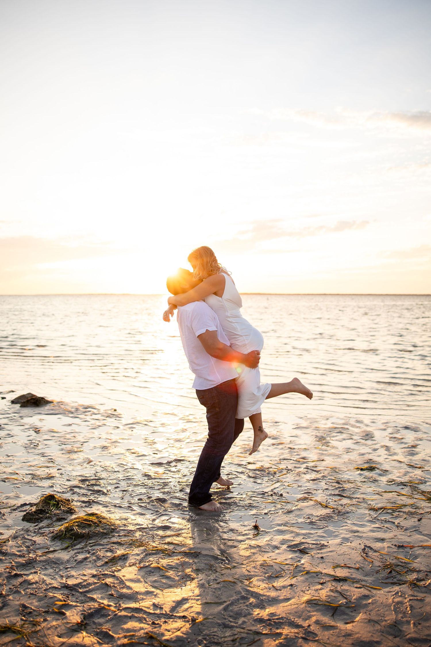 Couple embracing on a sandy beach at golden sunset, man lifting woman in romantic seaside moment.