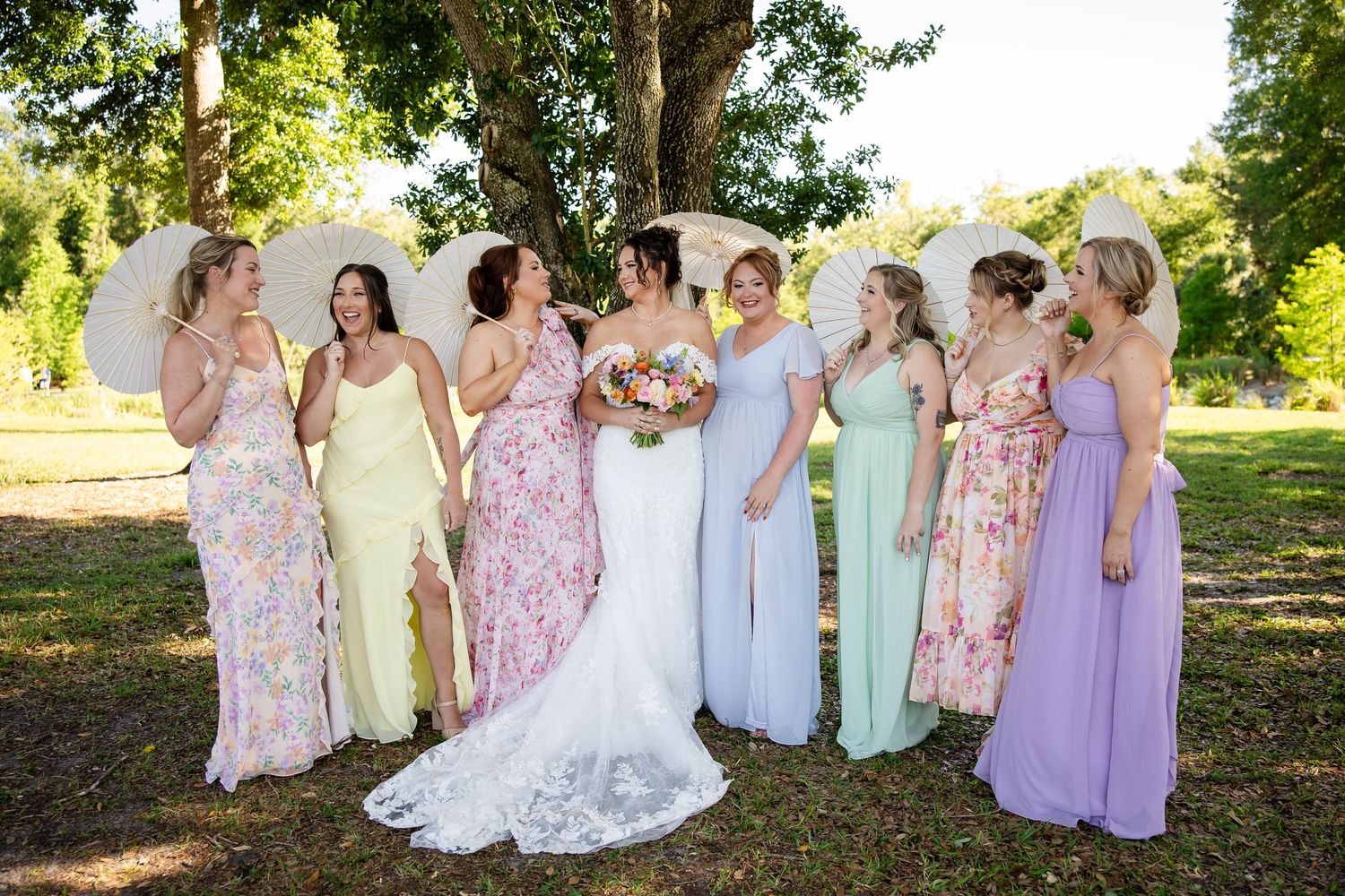 Bride in white lace gown poses with bridesmaids in colorful mismatched dresses holding parasols outdoors.