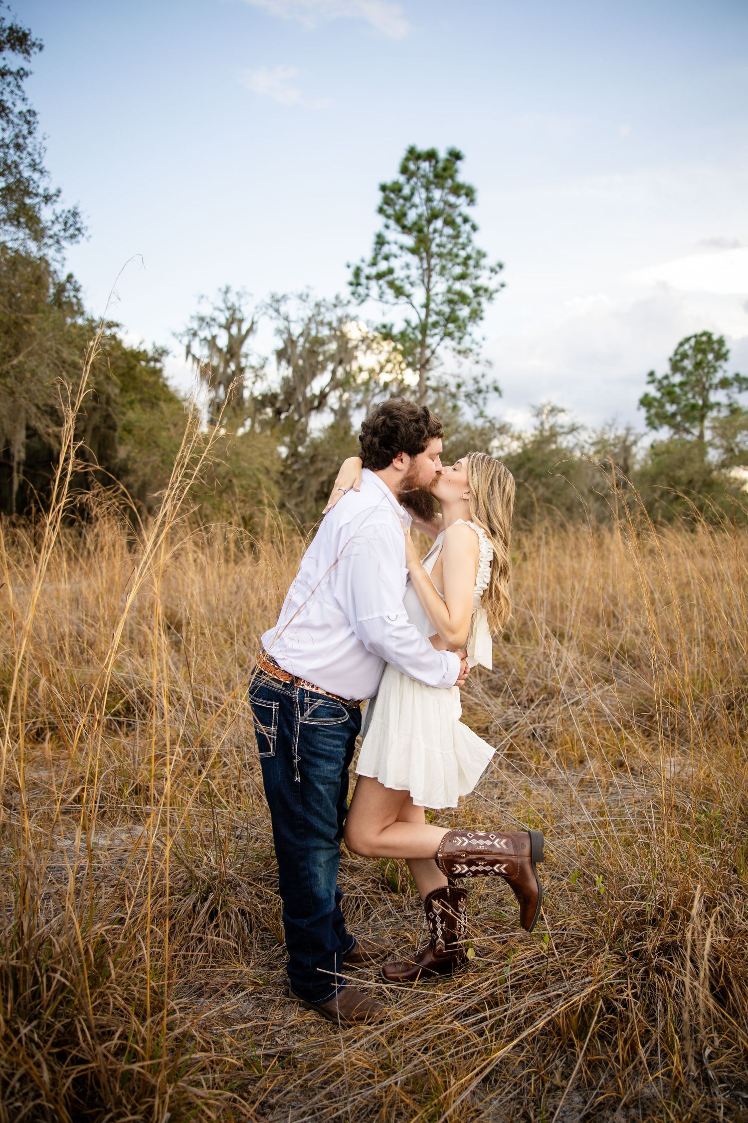 Couple sharing a romantic kiss in a golden field, man in white shirt and jeans, woman in white dress and cowboy boots.
