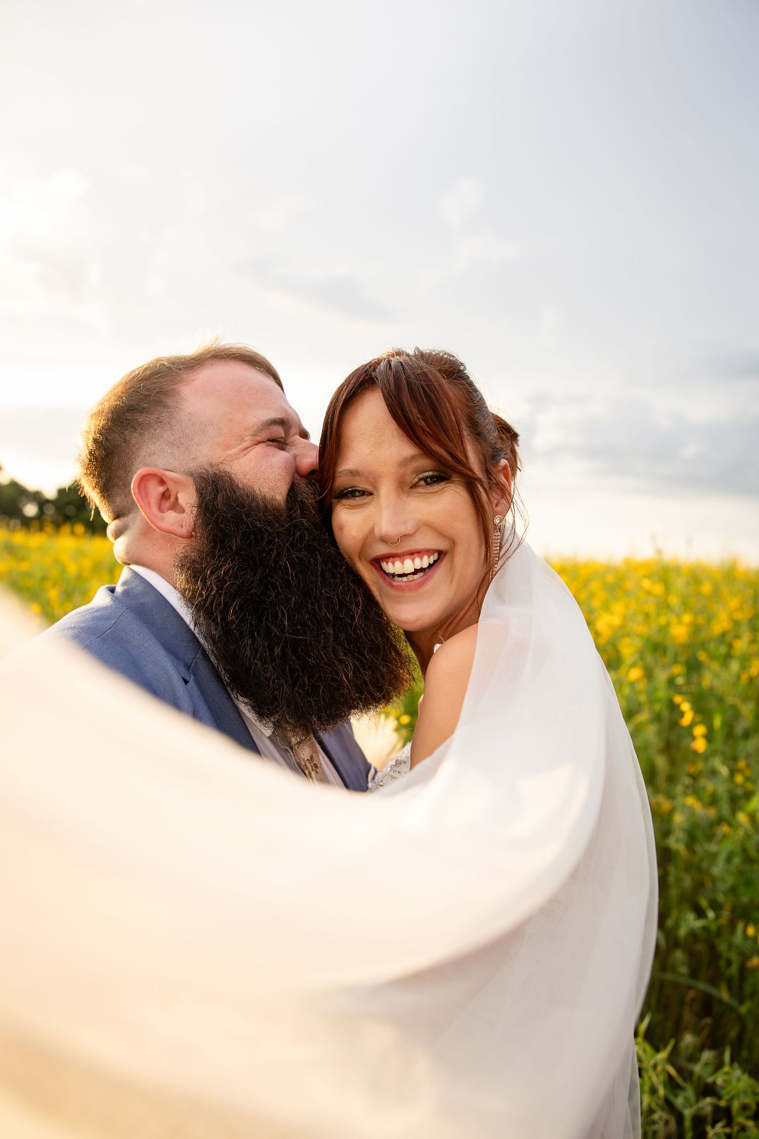 Bearded groom in blue suit kissing laughing bride in white veil in a yellow flower field at sunset.