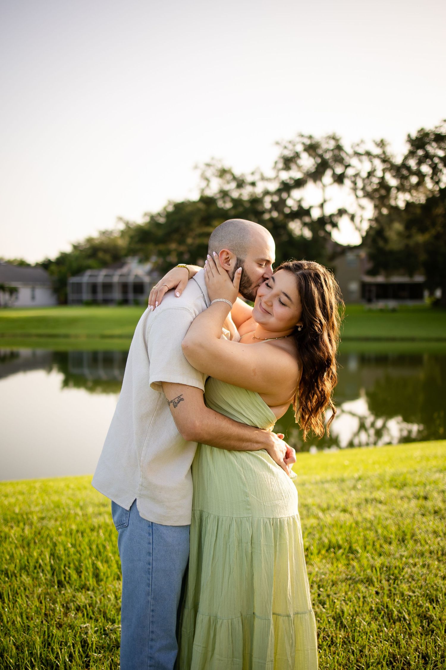 Romantic couple embraces by a serene lake at sunset, with lush grass and trees in the background.