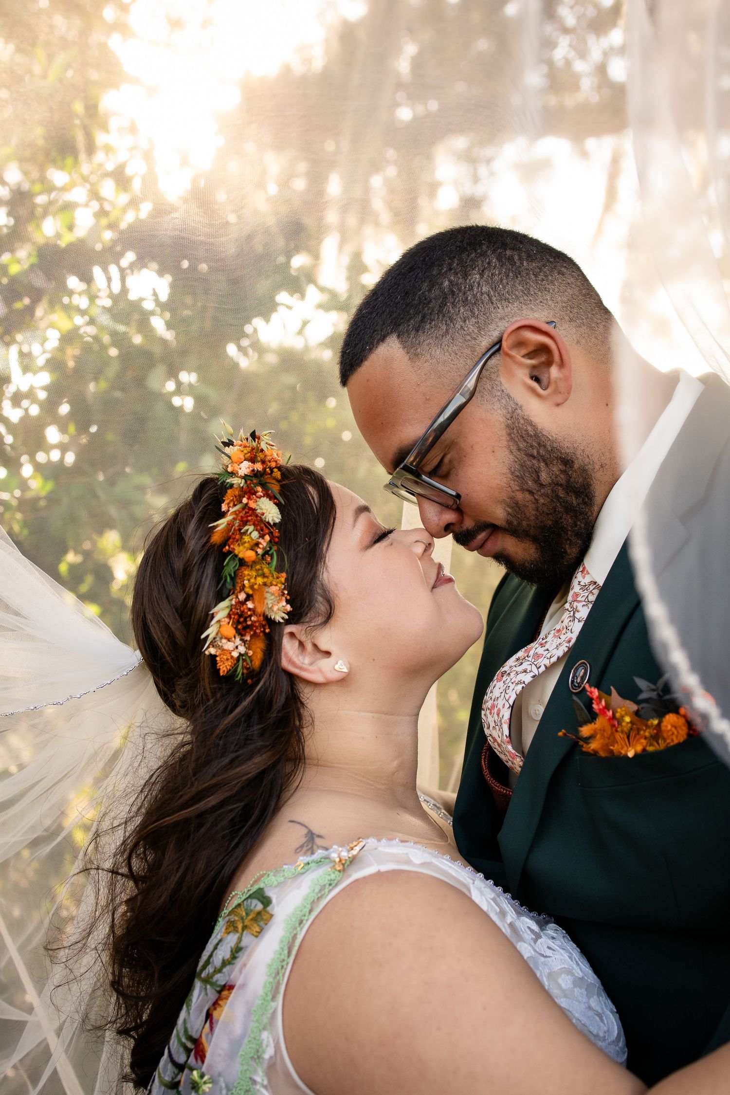 Romantic wedding couple shares tender moments in sunlit outdoor setting with autumn floral hair accessories.