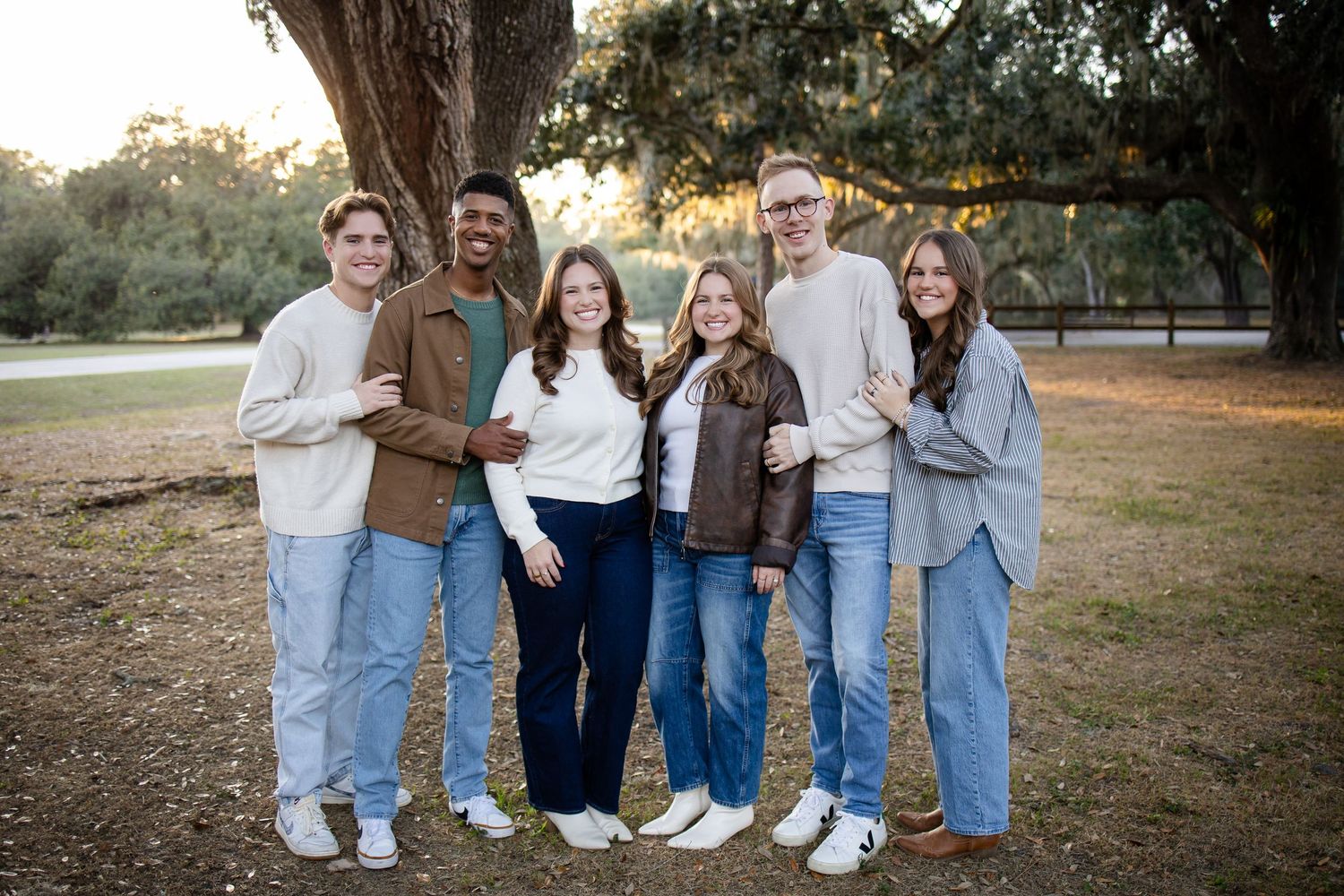 Group of friends in casual outfits posing together under a large oak tree at dusk.