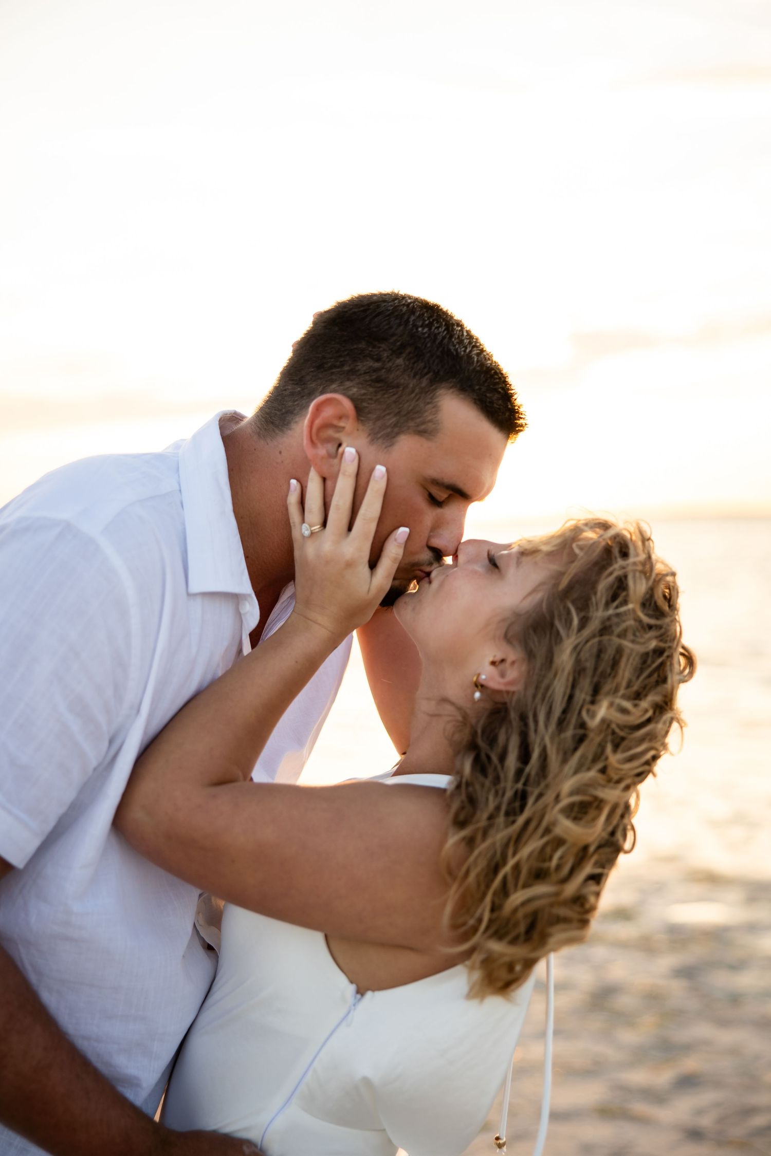 Couple sharing a romantic kiss on the beach at sunset, woman wearing white dress with curly blonde hair.