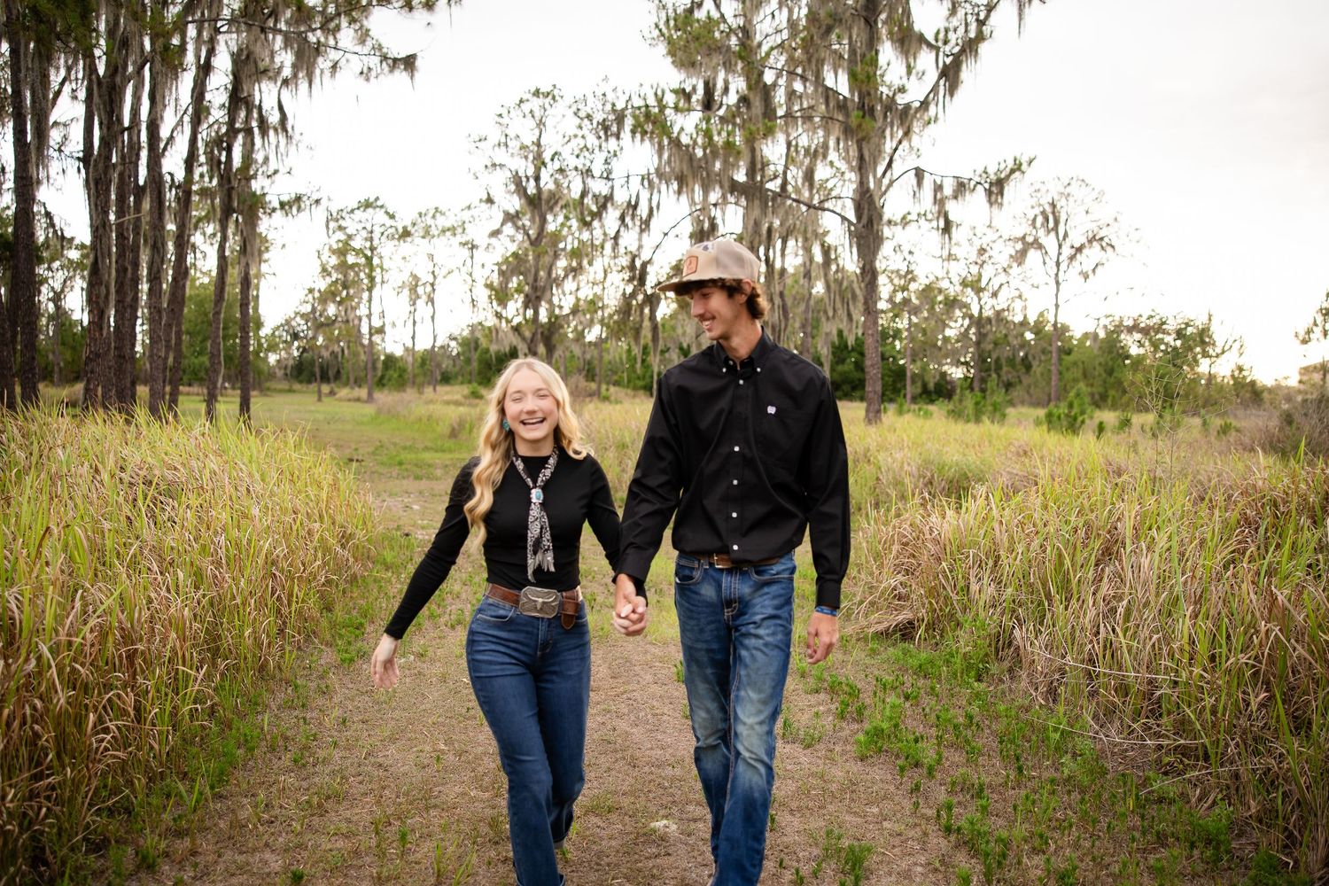Couple holding hands walking on a dirt path through tall grass and trees in a rustic outdoor setting.