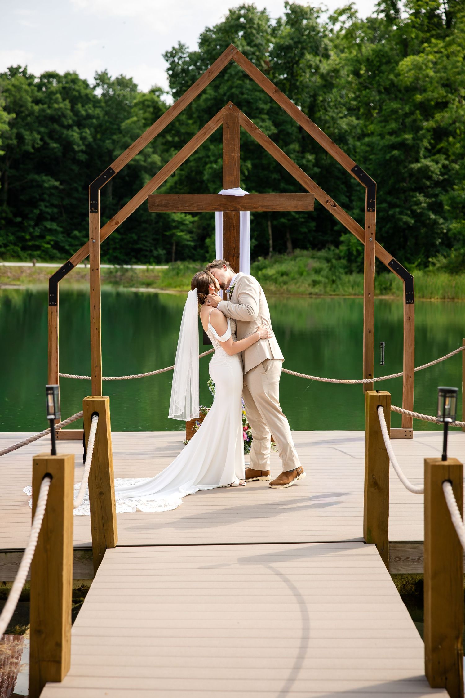 Bride and groom share first kiss under wooden arch on dock at scenic lakeside outdoor wedding ceremony.