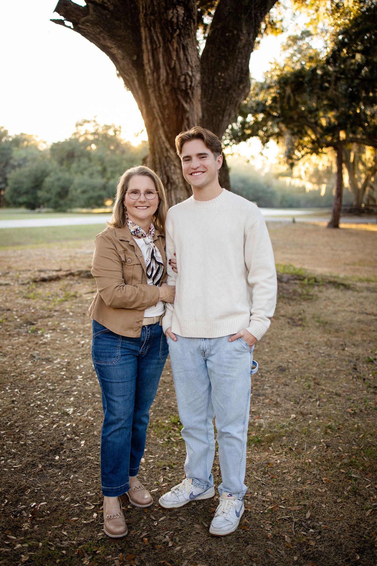 Two people in casual wear pose together under a large tree during sunset.