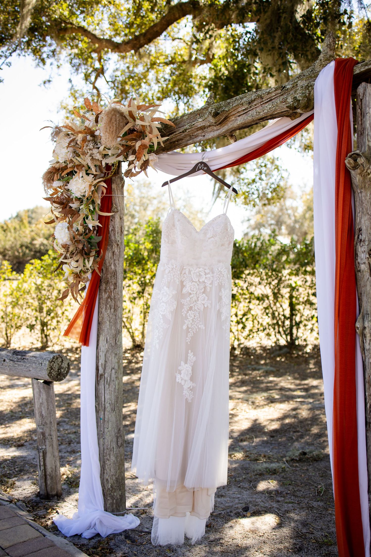 Wedding dresses and red fabric drape elegantly from rustic tree branches in an outdoor wedding setup.