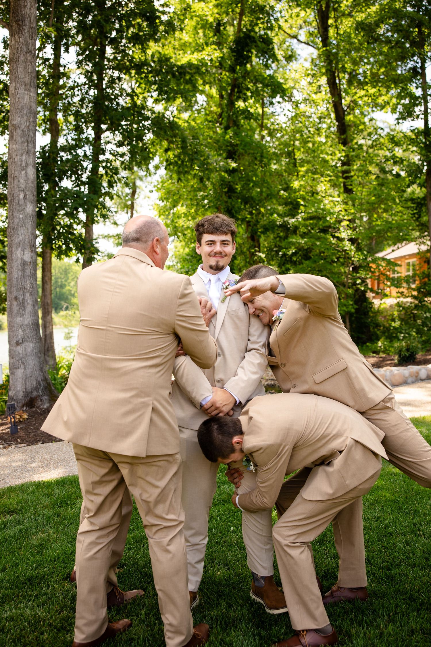 Groom in tan suit surrounded by groomsmen in matching suits posing playfully outdoors at a wedding venue.