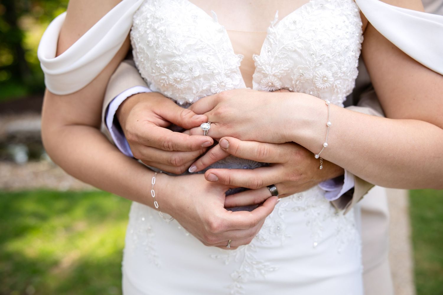 Bride and groom holding hands showing wedding rings, bride in white lace gown with diamond bracelet outdoors.
