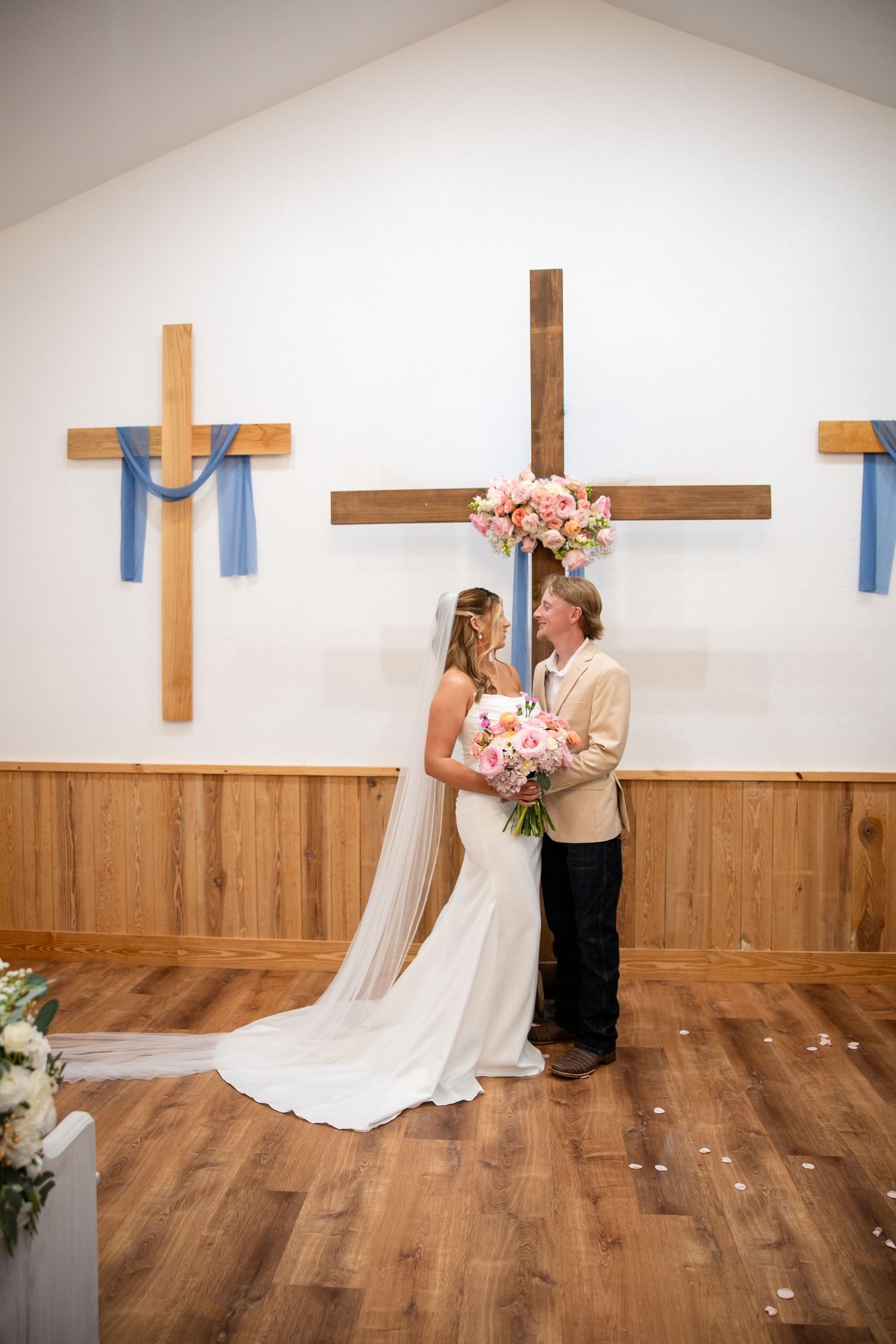 Bride and groom exchange vows in a rustic chapel with wooden crosses and blue ribbon decorations, bride holding pink bouquet.