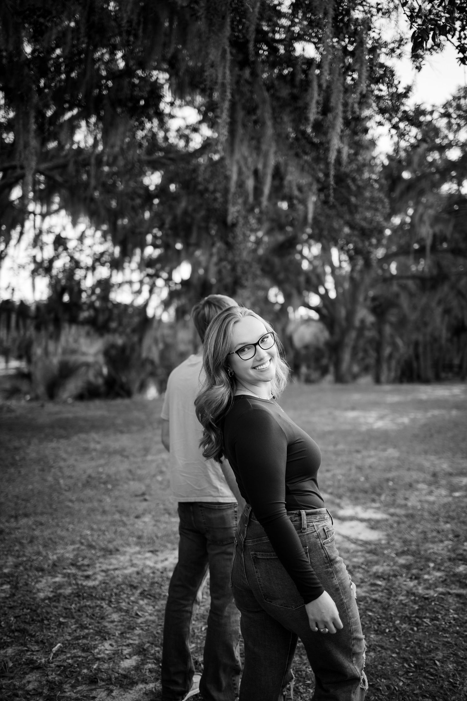 Black and white photo of a couple being playful outdoors under moss-draped trees.