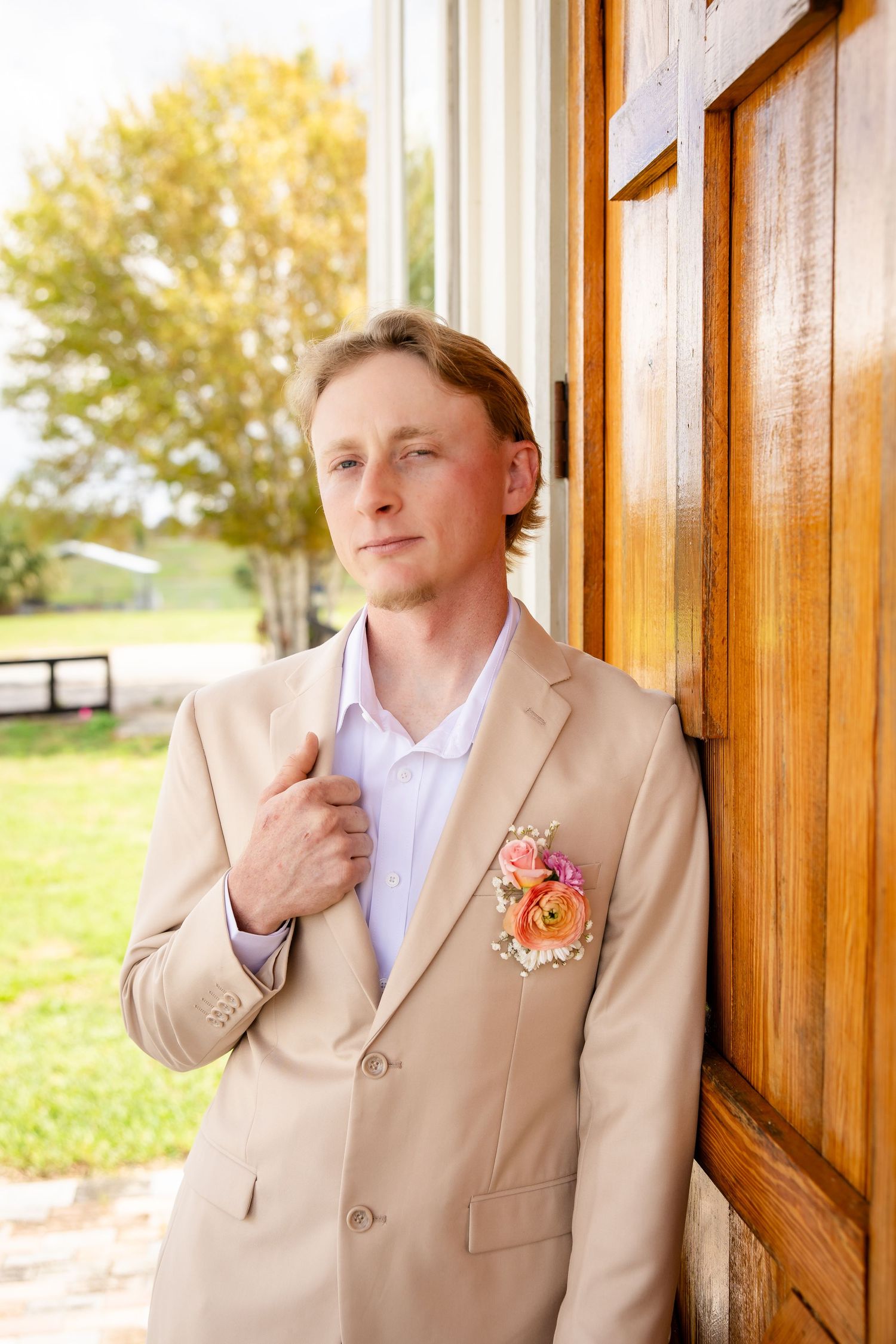 Groom in beige suit with floral boutonniere leans against wooden door outdoors on wedding day.