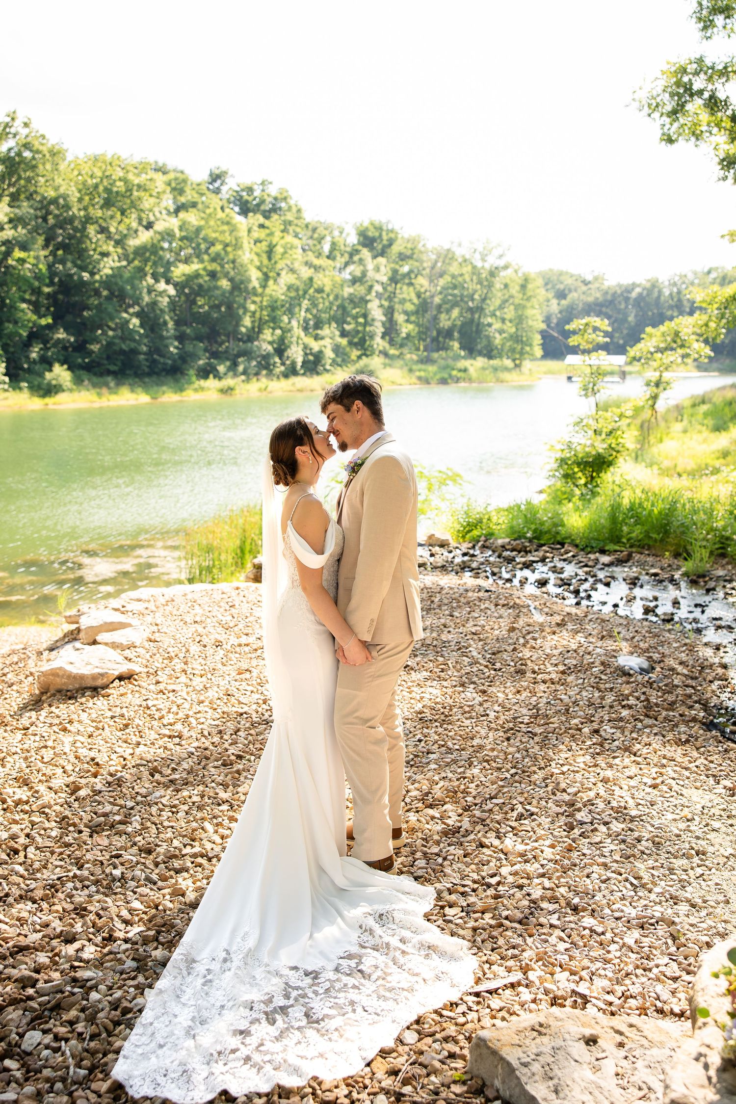 Bride and groom sharing an intimate moment by a sunlit river, bride in white lace gown, groom in tan suit.