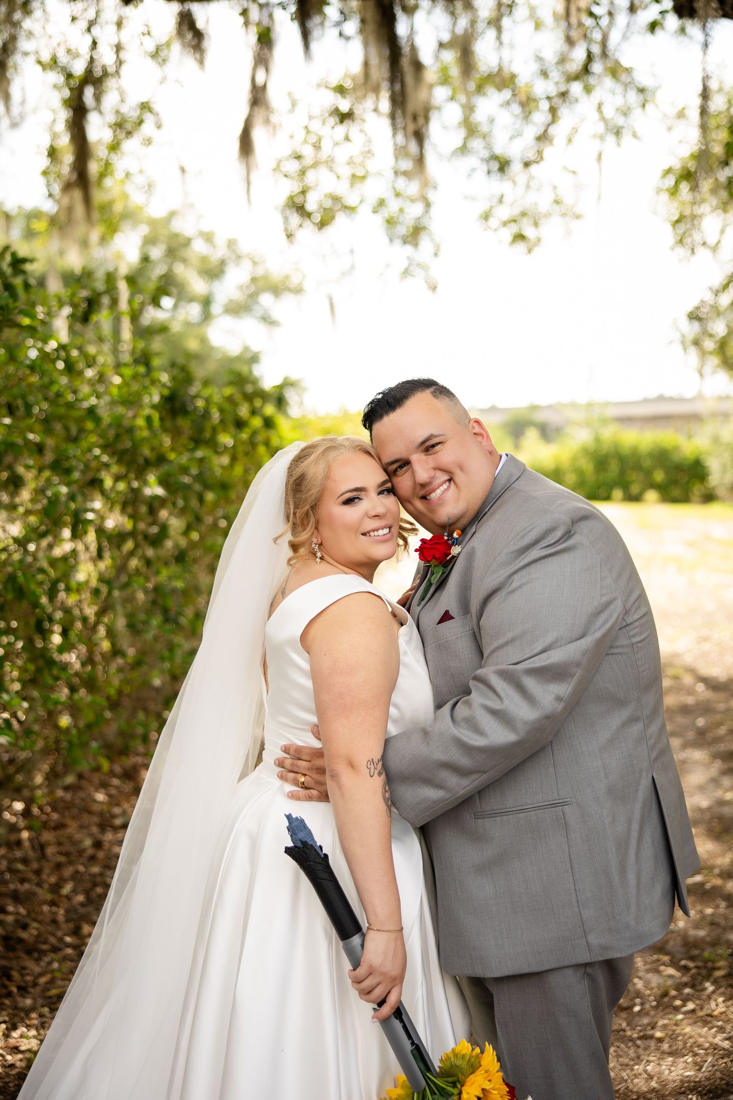 Bride in white gown and groom in gray suit embrace outdoors under Spanish moss trees, holding yellow bouquet.