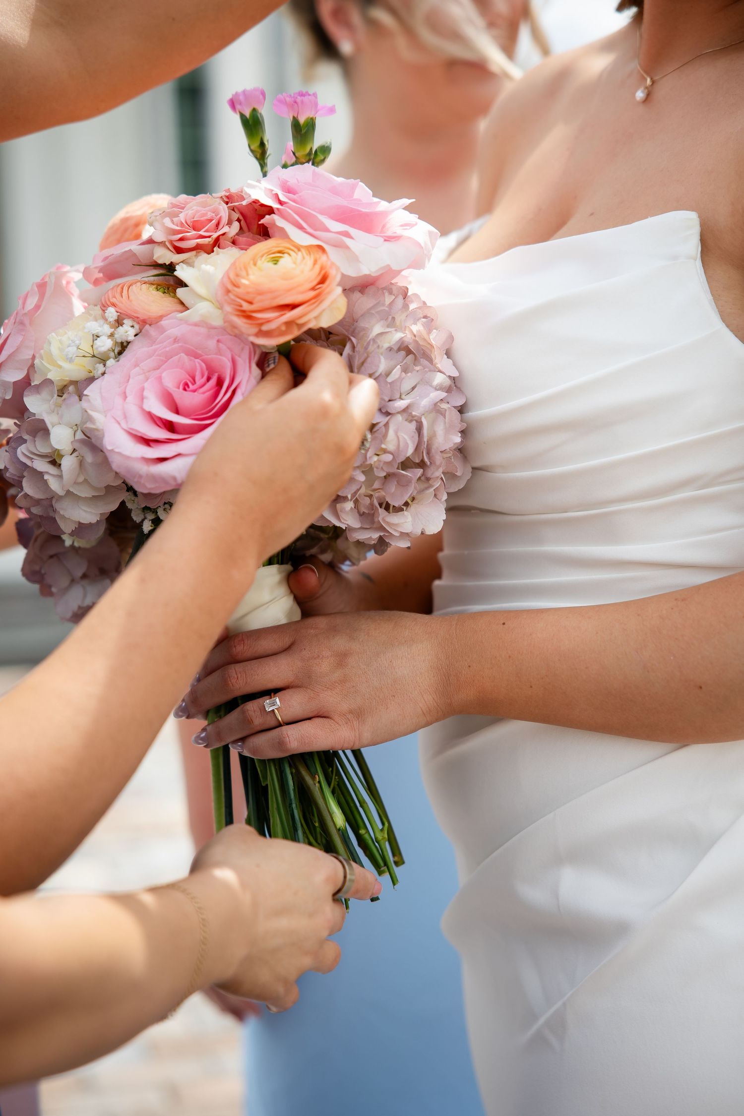 Bride in white gown holding pink and peach rose bouquet with hydrangeas, assisted by bridesmaid on wedding day.