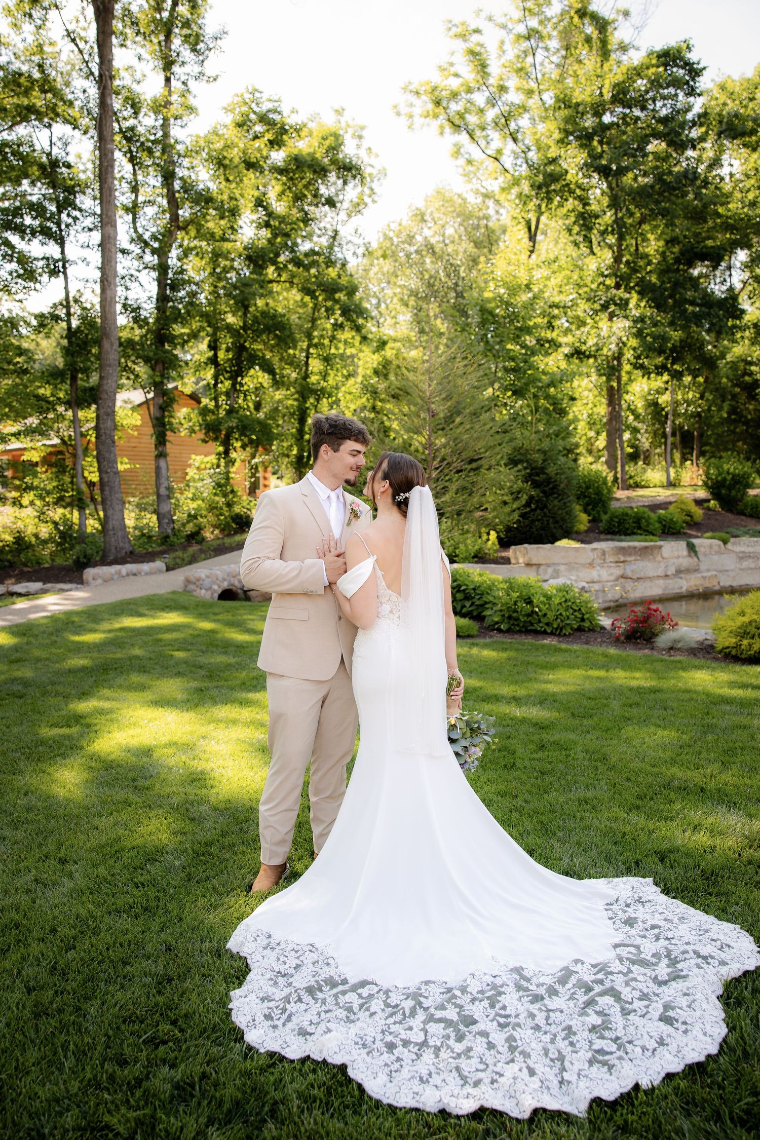 Bride and groom share a romantic moment on lush green lawn, her lace cathedral veil trailing elegantly behind her white gown.
