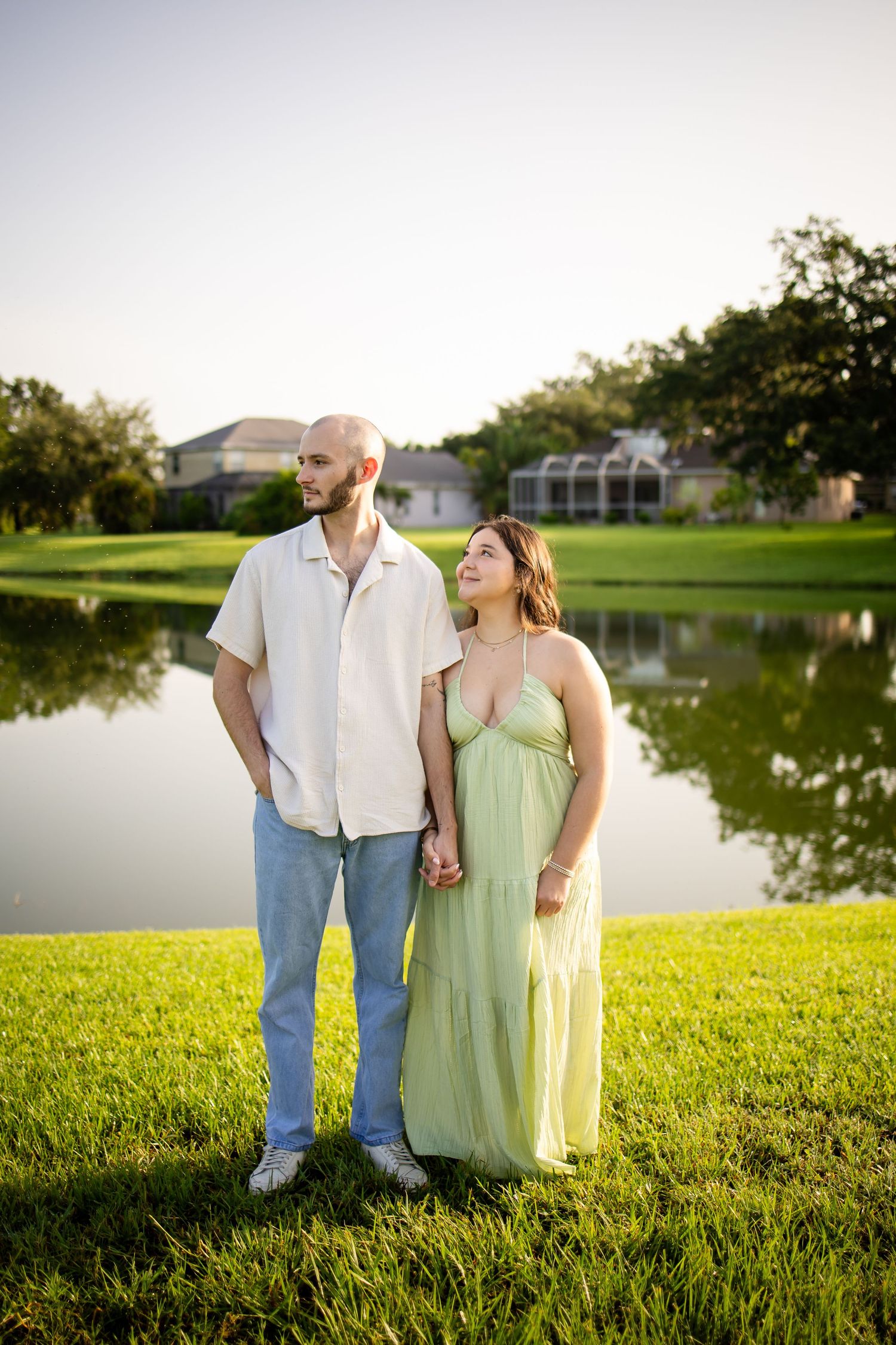 A couple holds hands while standing on green grass near a tranquil lake with houses in the background.
