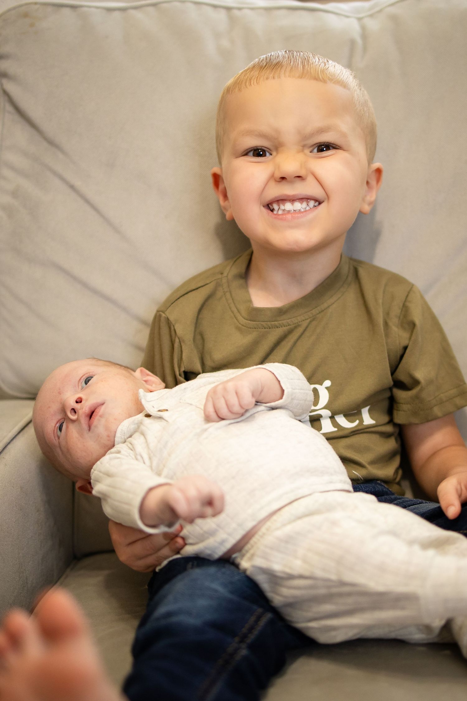 Two children sit together on a gray couch, with a young baby in a white onesie resting beside an older sibling in olive shirt.