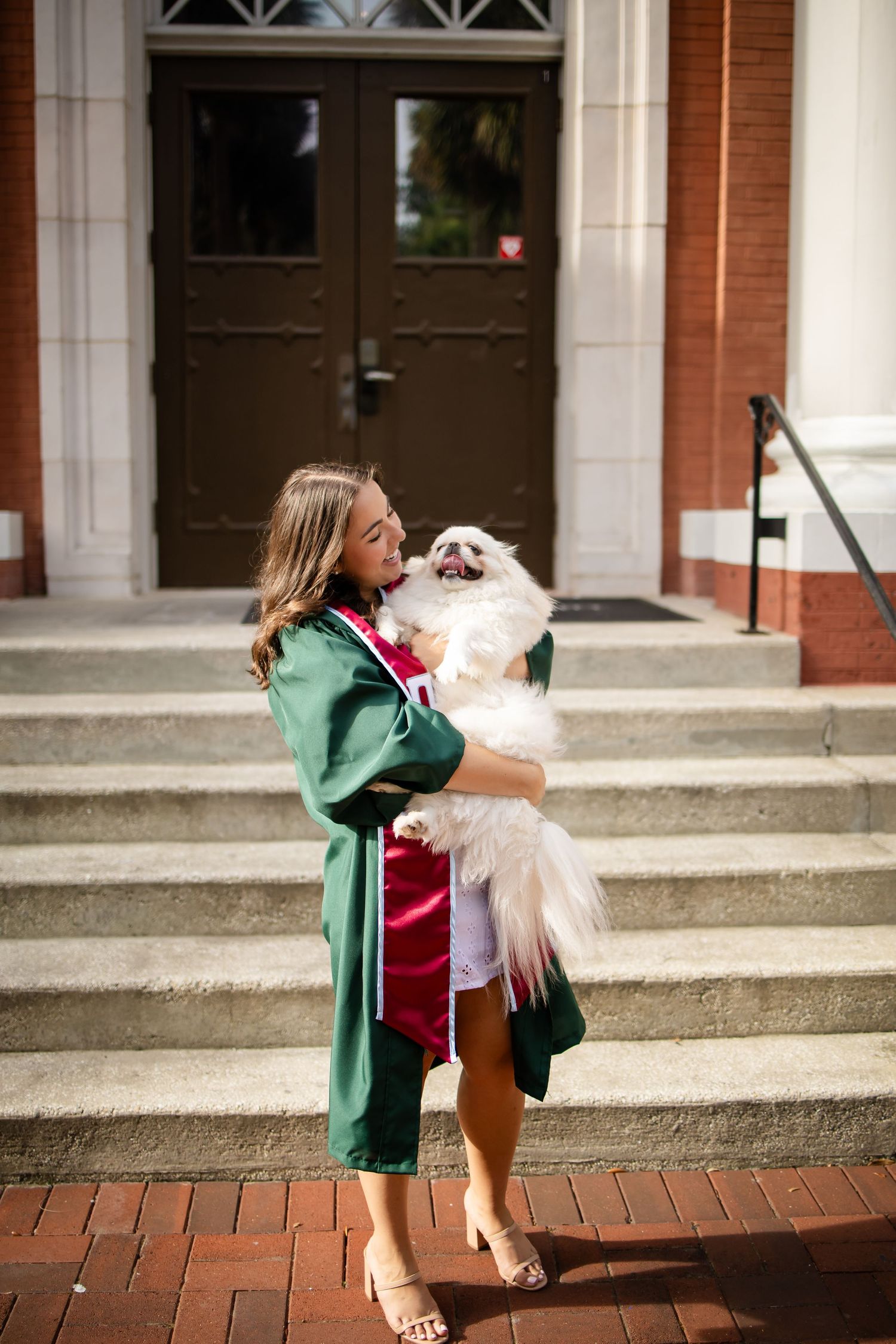 A graduate in green academic regalia holds a fluffy white dog while standing on brick steps outside building doors.