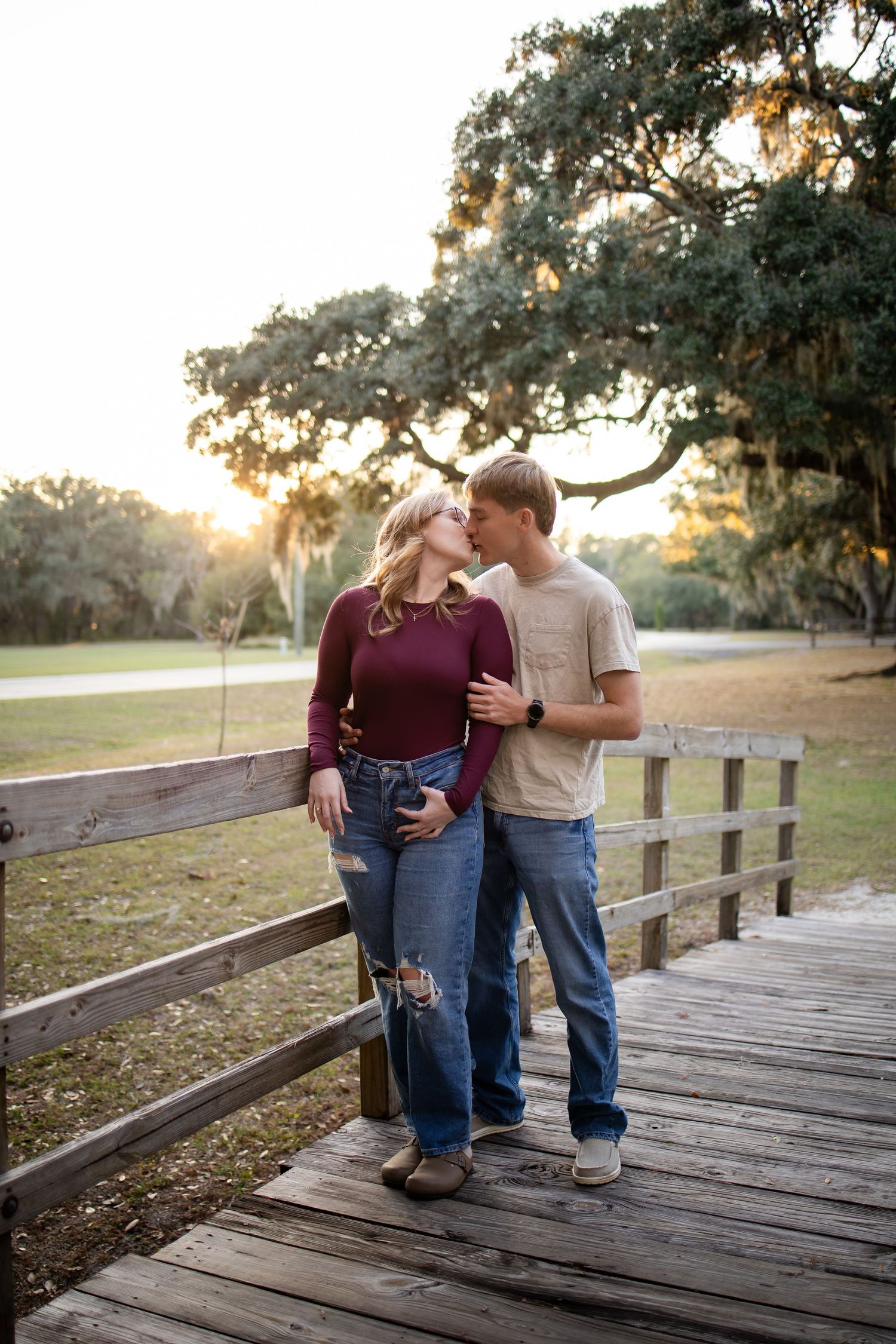 A couple shares an intimate moment on a wooden boardwalk at sunset surrounded by oak trees.